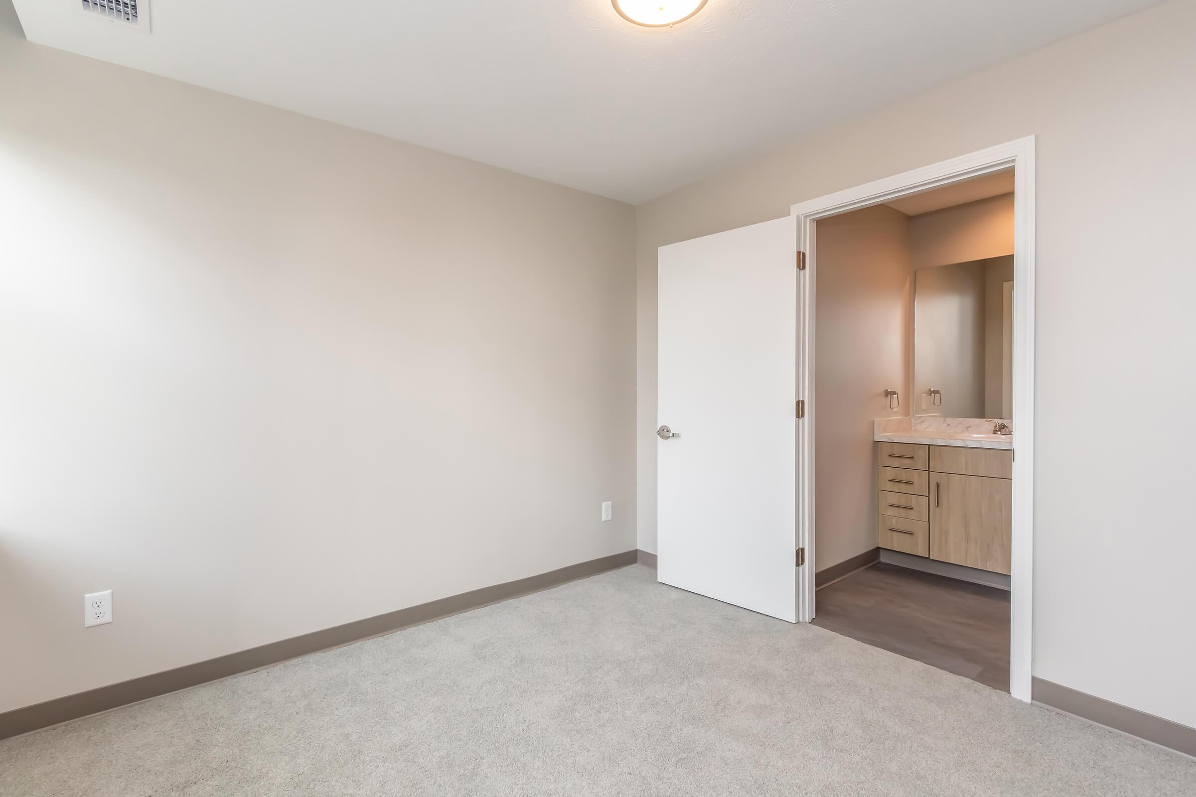 A vacant room with light beige walls and a soft carpet floor. A white door leads to a bathroom with wooden cabinetry and a mirror. Natural light fills the space, creating a bright and airy atmosphere. The room is sparsely decorated, emphasizing its potential for personal touches.