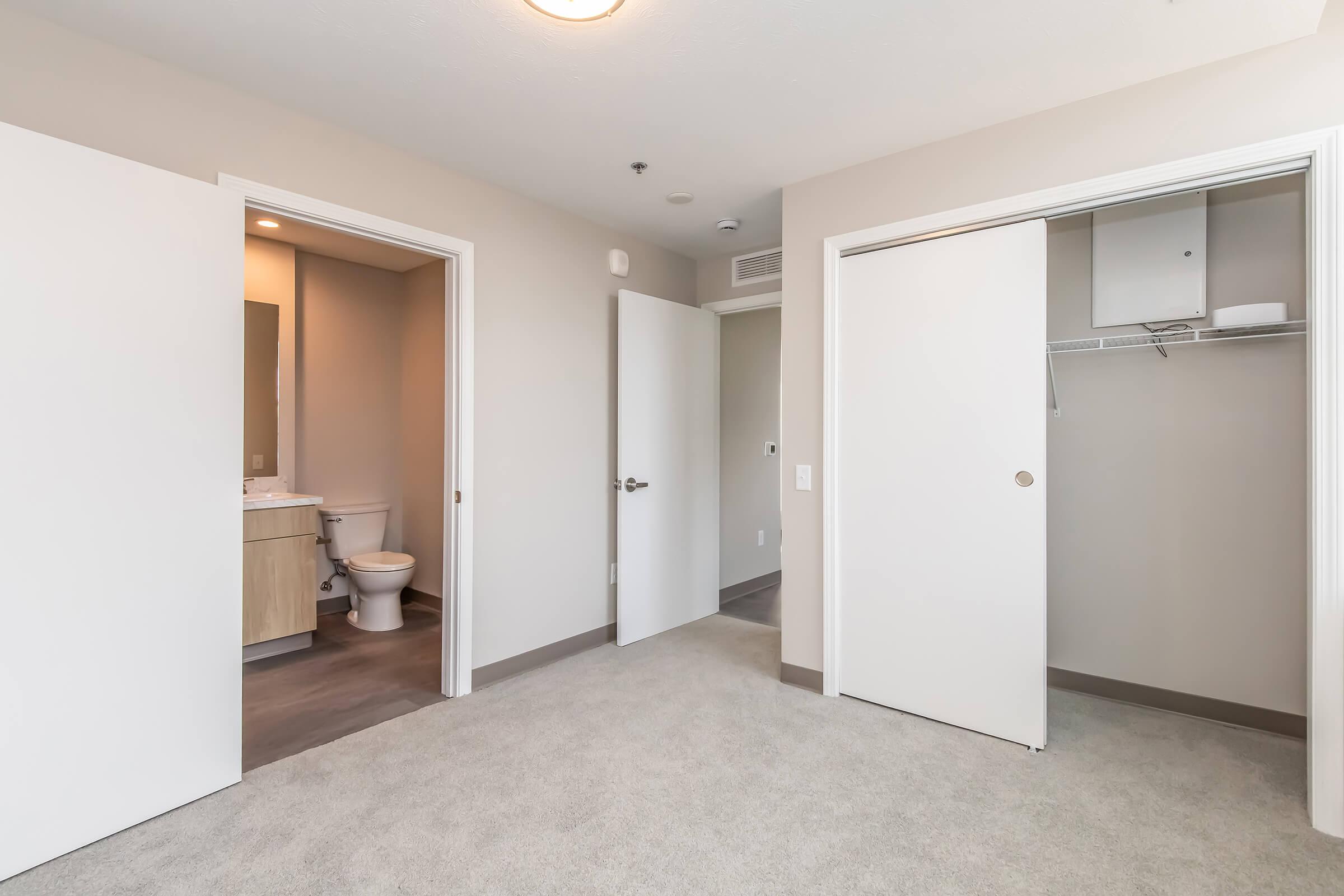 A modern, empty room featuring light-colored walls and carpet. Two open doors lead to a bathroom and a closet. The bathroom has a simple vanity and toilet visible. The overall ambiance is clean and minimalist, suitable for a living space.