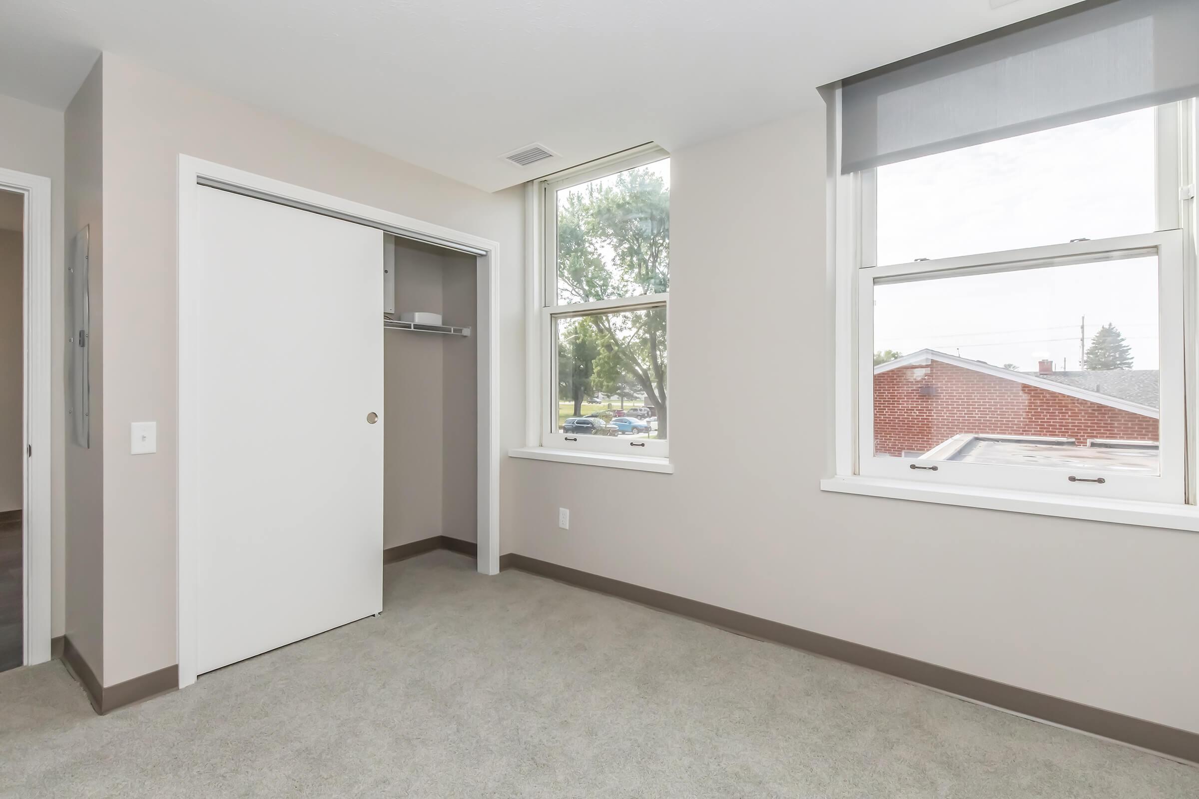 A well-lit, empty bedroom featuring light-colored walls and carpet. There is a closet with a white sliding door and two windows allowing natural light. The view outside includes a glimpse of a street and nearby buildings. The room has a modern and clean appearance.