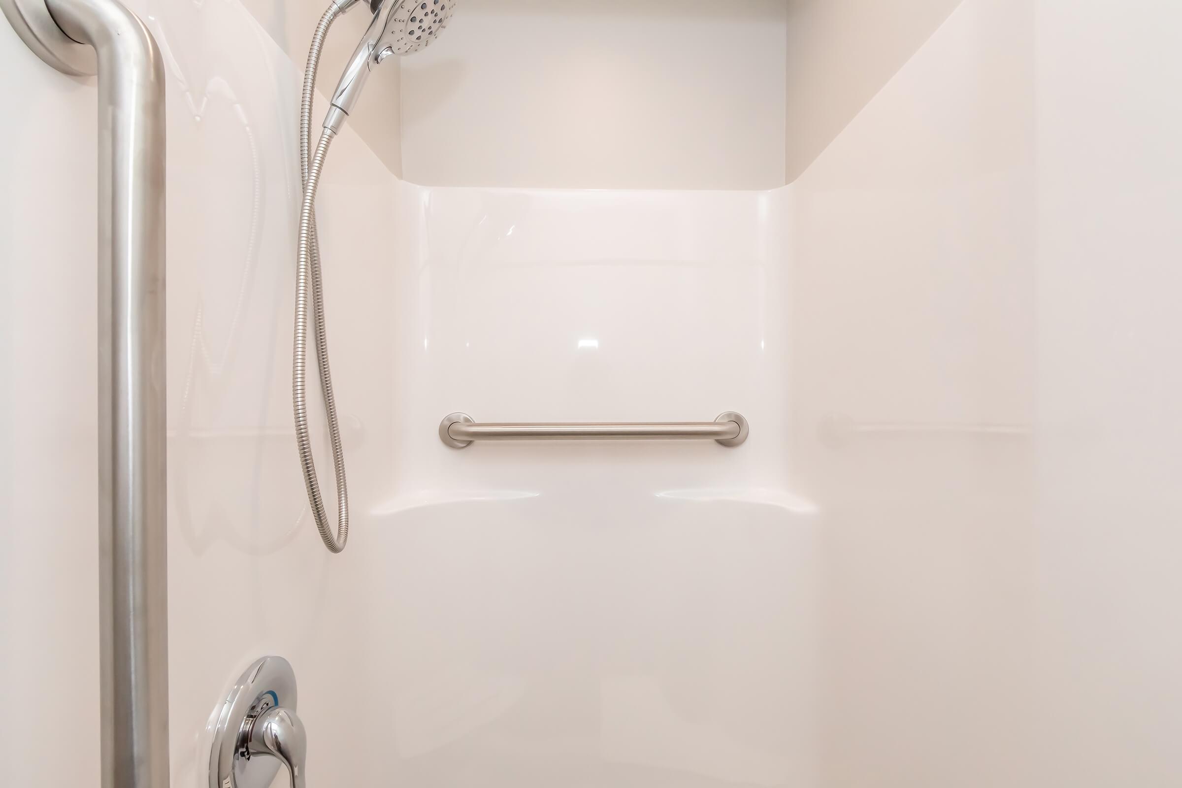 A clean, modern shower stall featuring smooth white walls, a handheld showerhead with a flexible hose, and a stainless steel grab bar installed on the wall for safety. The space is well-lit, creating a bright and airy atmosphere.