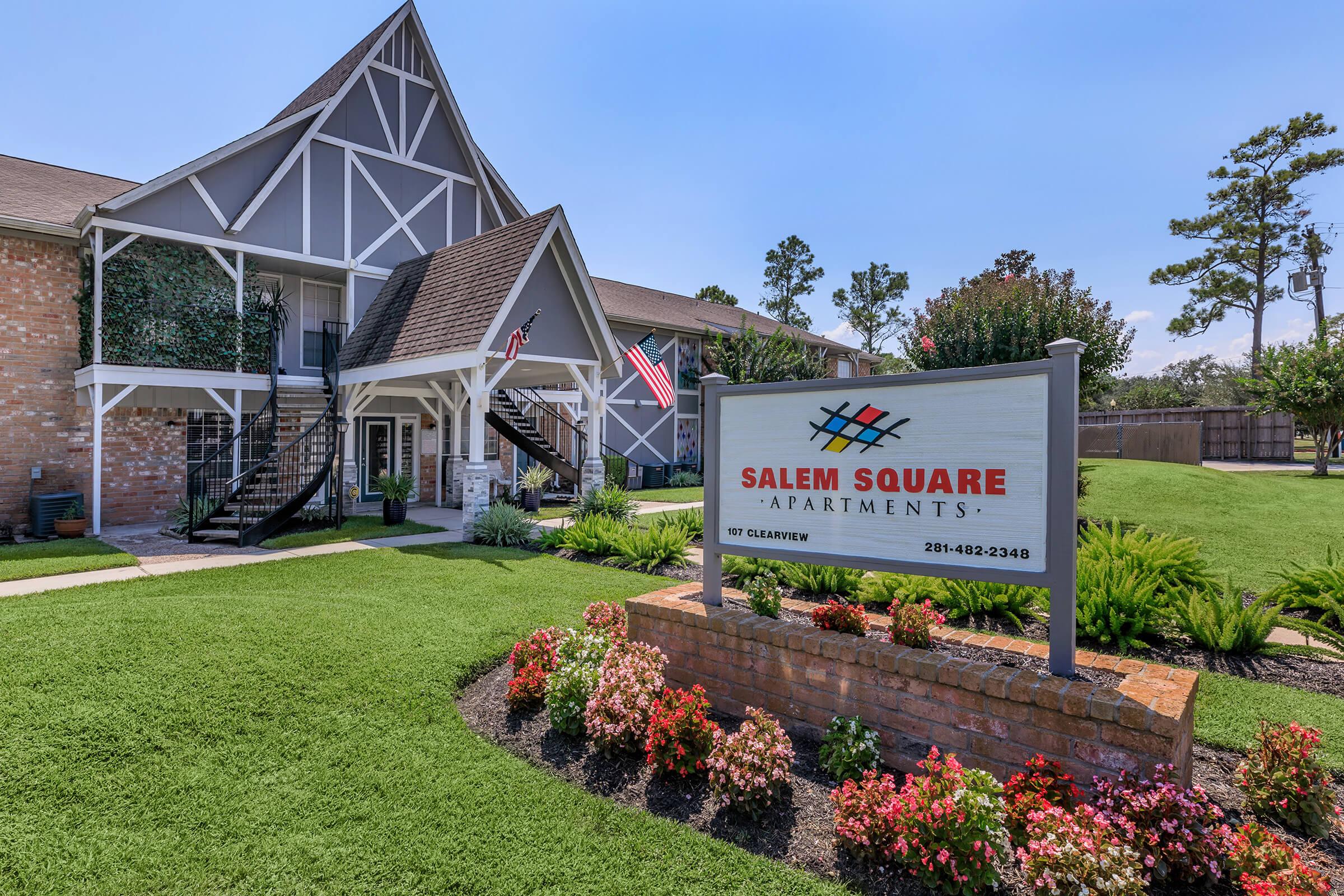 A view of Salem Square Apartments, featuring a multi-story building with a peaked roof and balconies. A prominent sign for the apartments is displayed in front, surrounded by colorful flower beds and well-maintained green lawns. American flags are visible on the building. Clear blue skies overhead.
