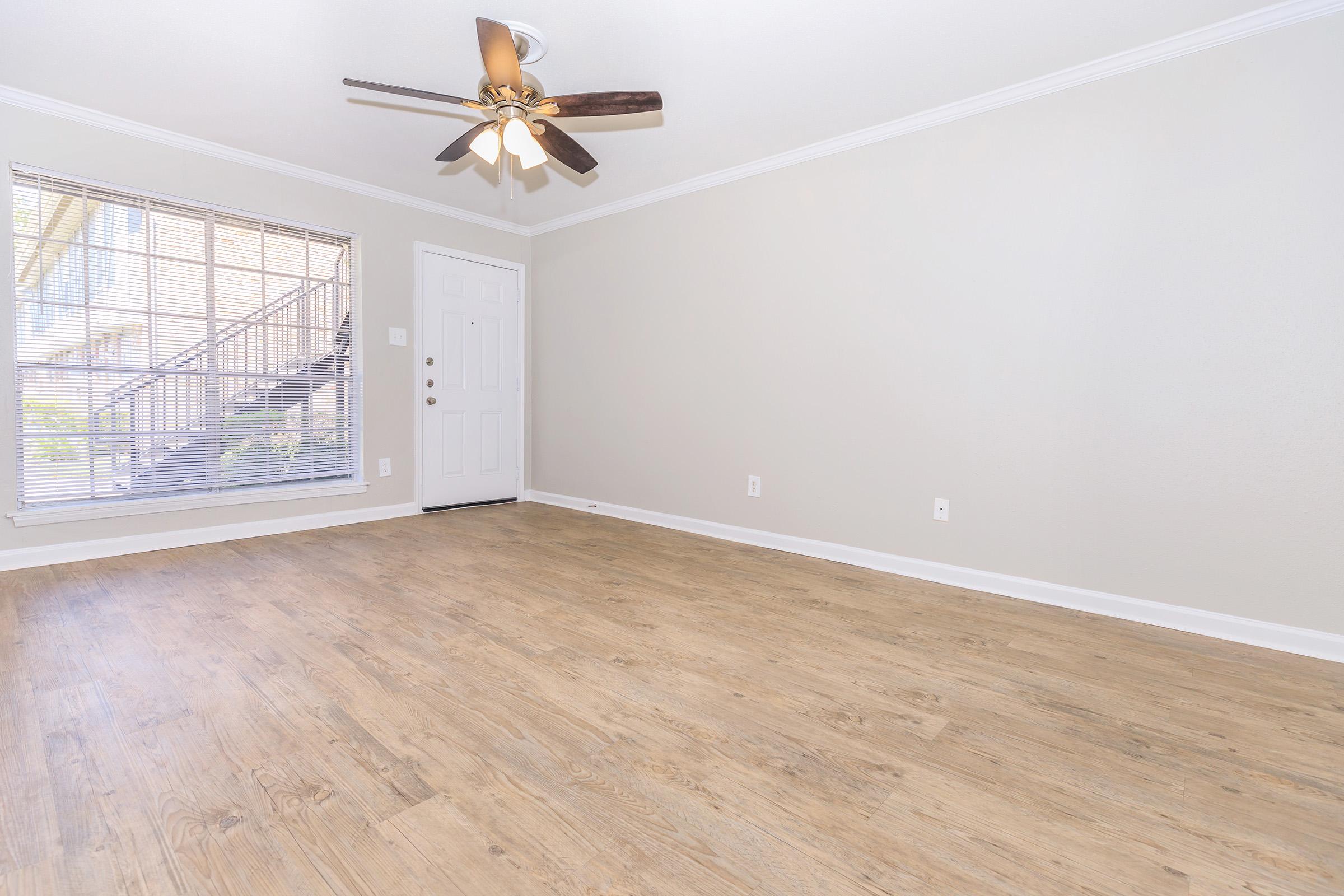Spacious interior of an empty room featuring a light-colored wall, wooden flooring, a ceiling fan, and a window with blinds. A door is visible on the right, leading outside, and there is a subtle natural light illuminating the space.