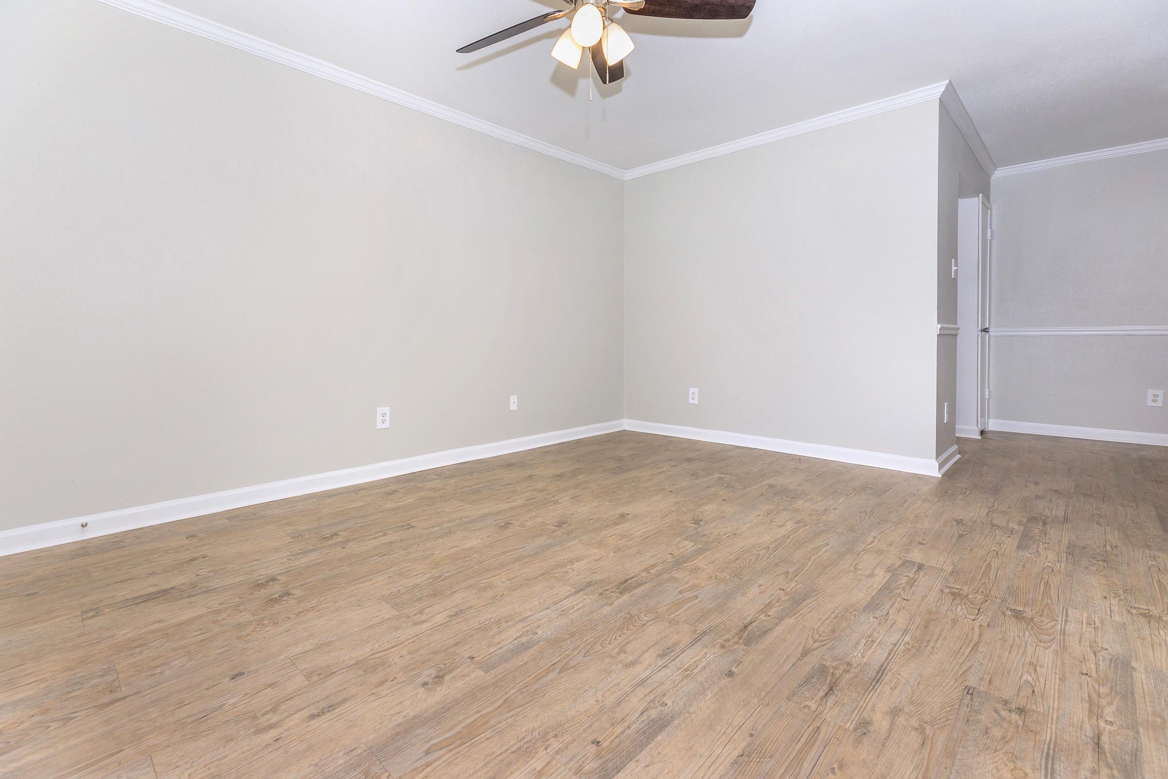 A spacious, empty living room featuring light-colored walls and new wooden flooring. A ceiling fan hangs above, providing a modern touch. The room has clean lines and an open layout, ideal for decorating according to personal taste.
