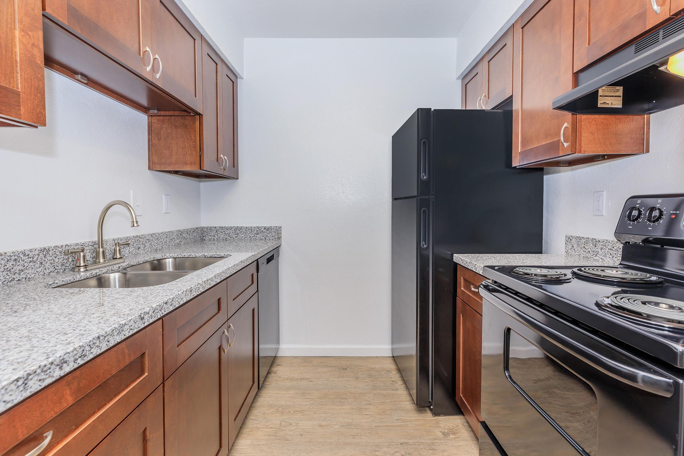 A modern kitchen featuring dark wood cabinets, a black refrigerator, and a black stove. The countertops are made of granite, and there are stainless steel sinks with two basins. The flooring is light-colored, giving the space a clean and contemporary look.