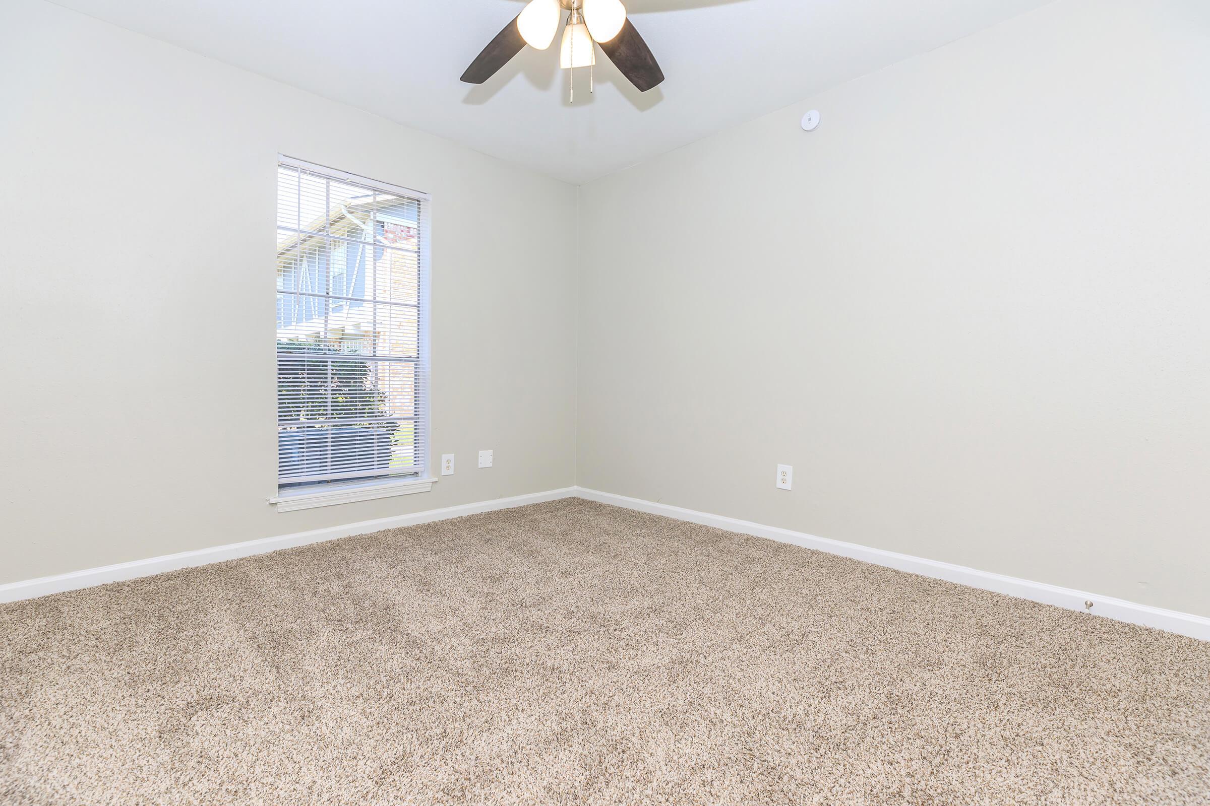 A vacant room with light beige walls and a ceiling fan. The floor is covered with soft, light brown carpet. A window with white blinds allows natural light to enter, and a small outdoor view is visible through the window. The space is clean and uncluttered, perfect for personalization.