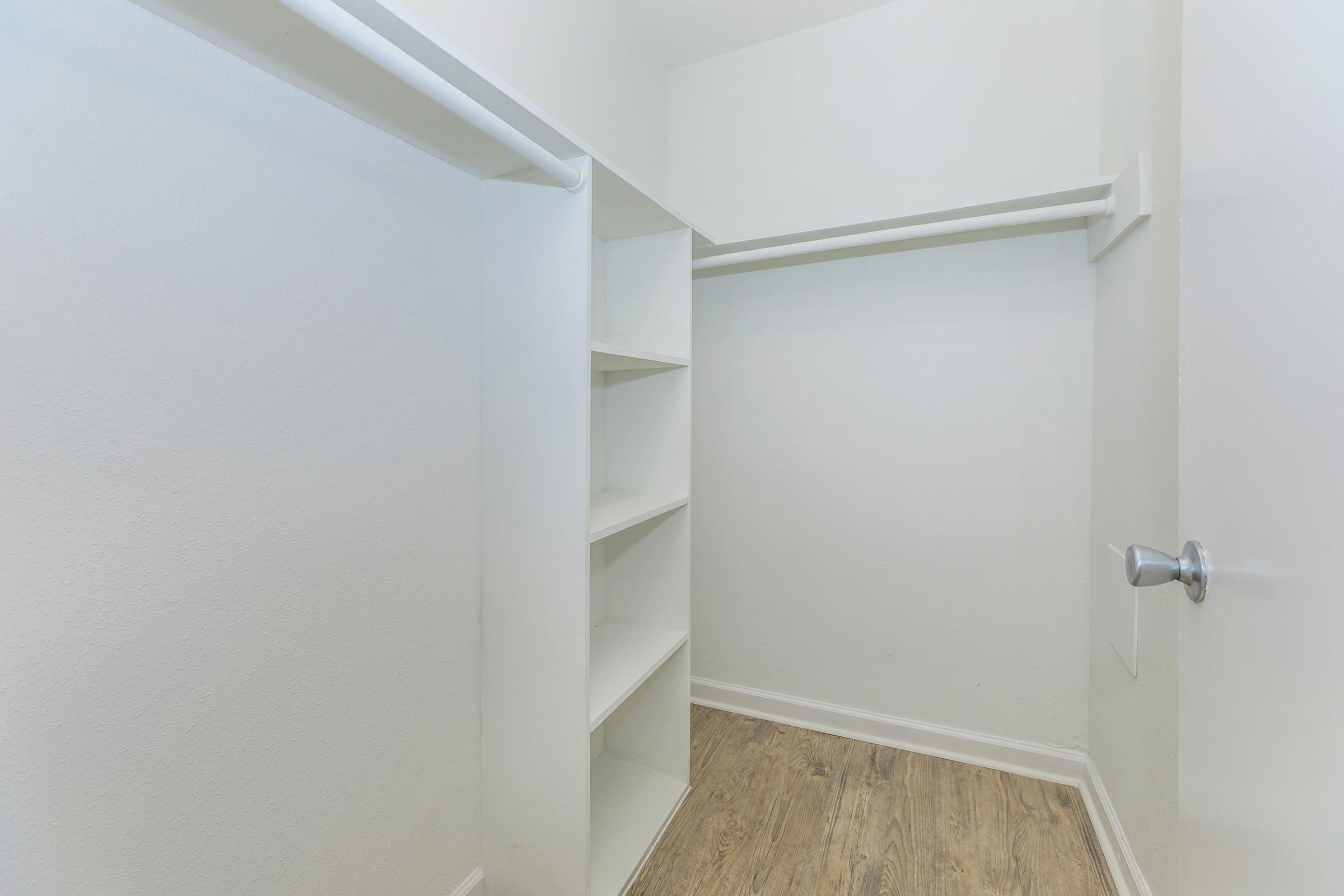 Empty walk-in closet featuring white walls, a single hanging rod, and built-in shelves on one side. The floor has a light wood finish, and a door is visible at the right side of the image, indicating the entrance to the closet.