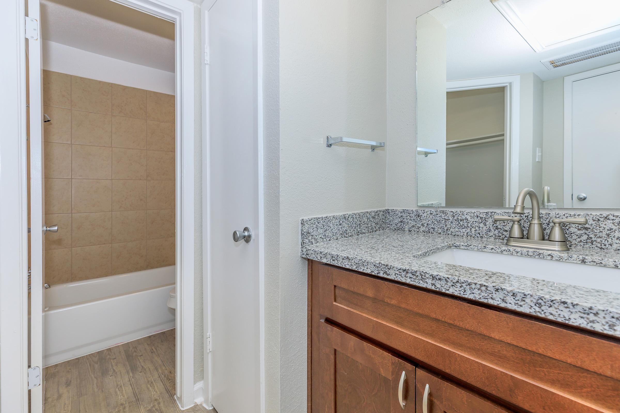 A bathroom featuring a granite countertop with a sink and modern fixtures. In the background, a beige-tiled area is visible, leading to a bathtub. The walls are painted light gray, and there are two doors: one entering the bathroom and another leading to a separate space. Natural light brightens the area.
