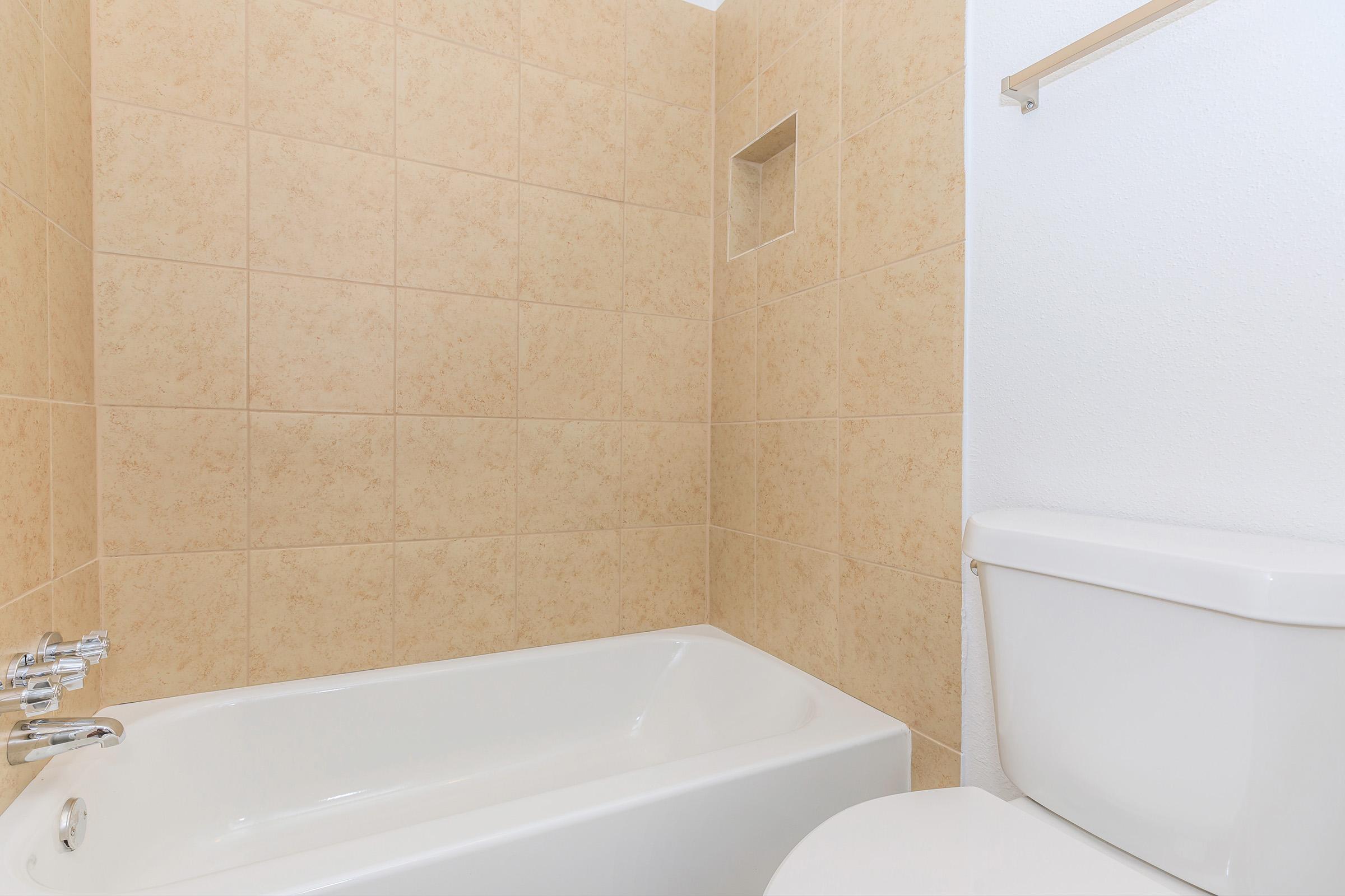 A clean, modern bathroom featuring a beige tiled wall, a white bathtub, and a white toilet. The bathtub is positioned against the wall, with a built-in shelf niche visible in the tile. The overall design is simple and contemporary, emphasizing a light and spacious feel.