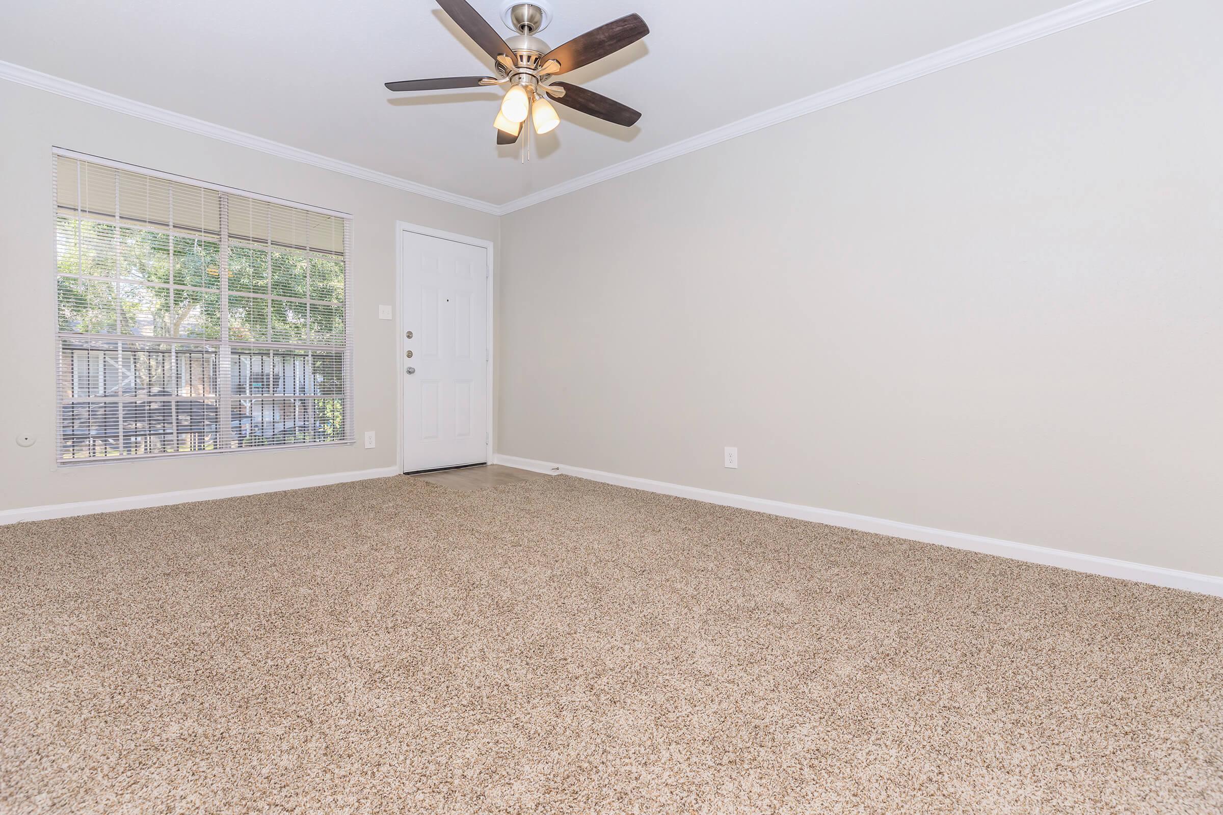 A spacious living room with light beige carpet, minimalist decor, and a ceiling fan with four light bulbs. A window allows natural light to enter, and there is a door leading outside. The walls are painted light gray, creating a calm and inviting atmosphere.