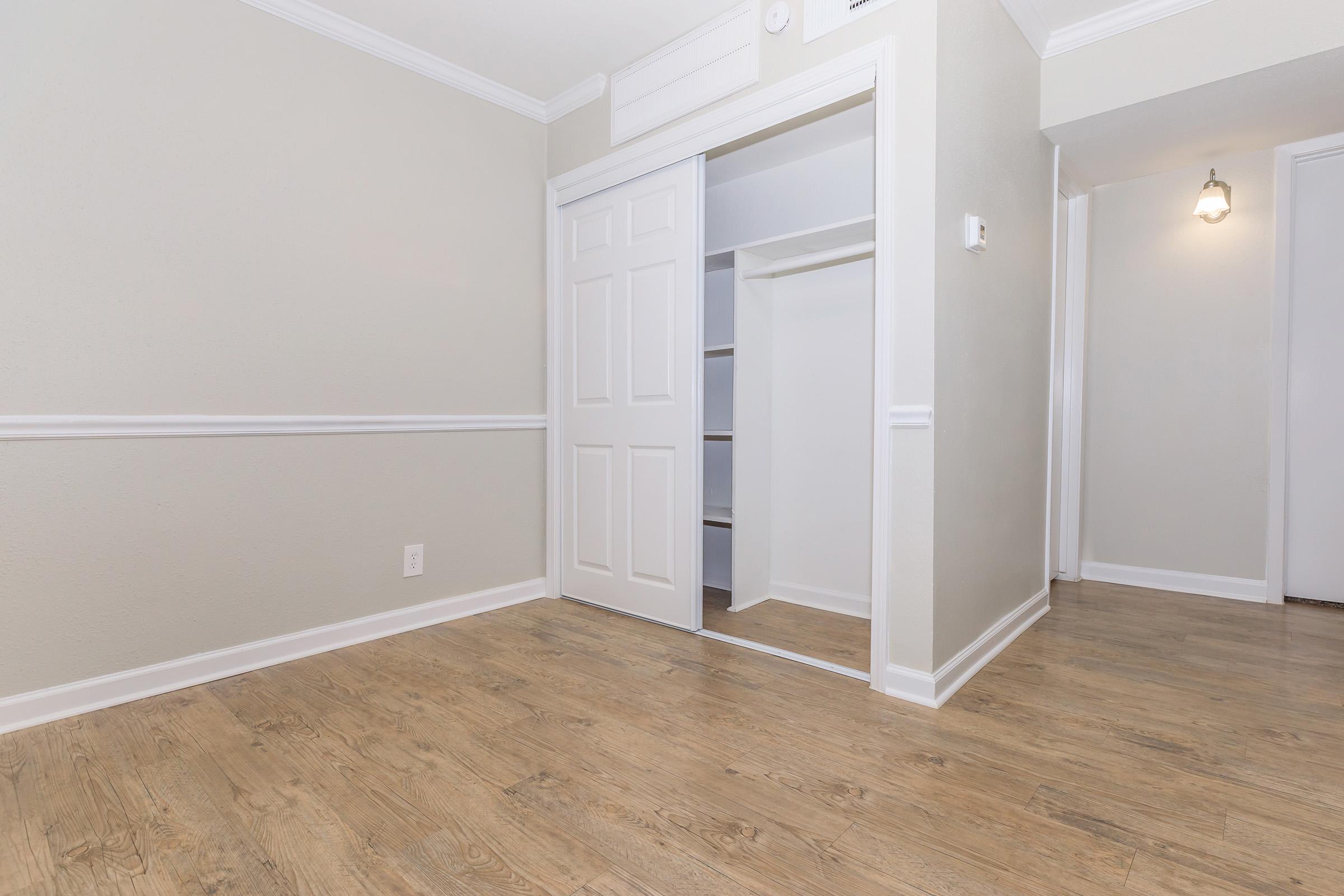 A spacious room featuring light gray walls and a wooden floor. There's a double-door closet on one side with shelving visible inside. The room includes a white baseboard and crown molding, with a wall-mounted light fixture. The opposite wall shows a doorway leading to another area.