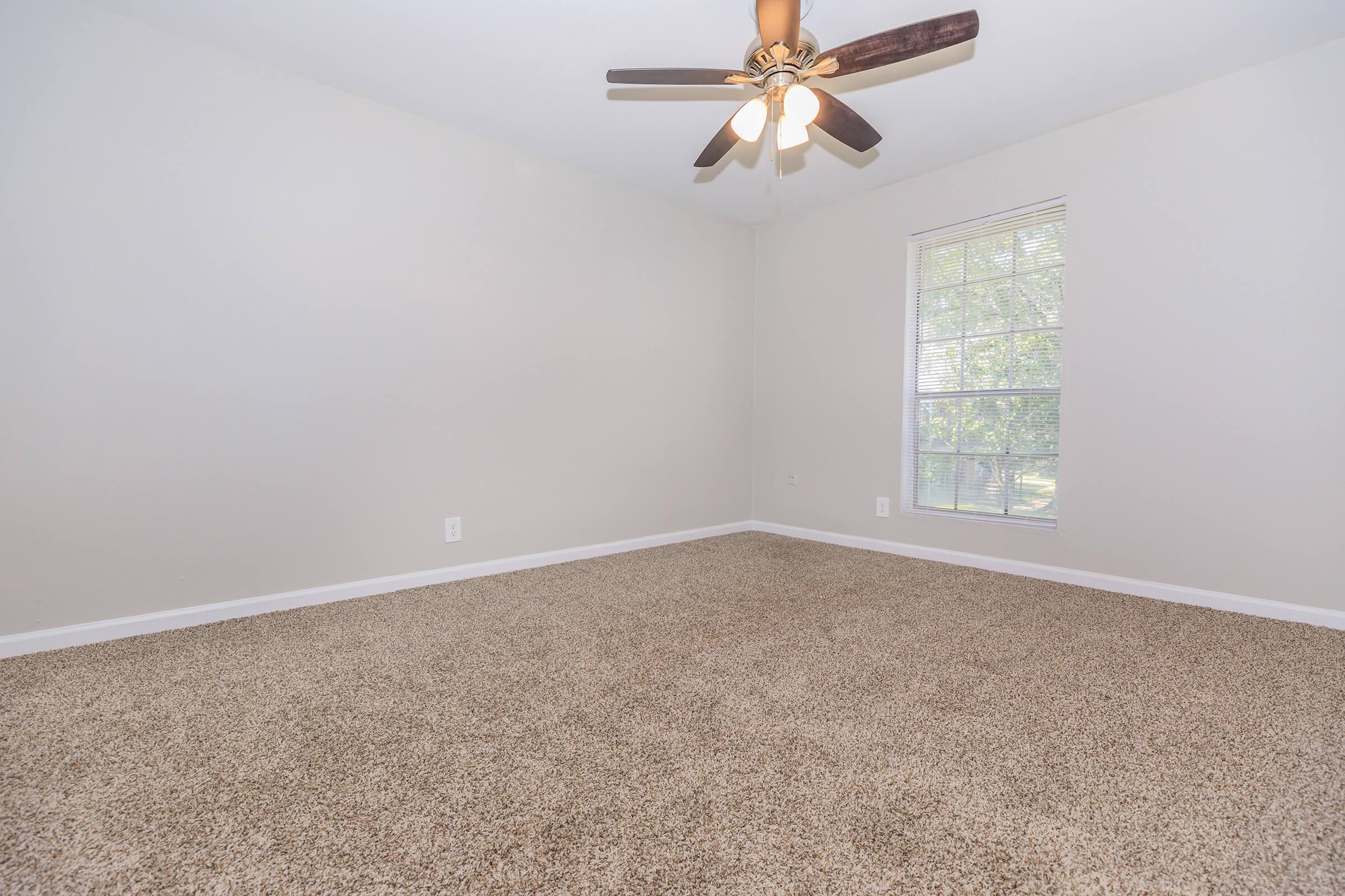 A light-colored room with beige carpet, featuring a ceiling fan with wooden blades. One window allows natural light, revealing greenery outside. The walls are painted a soft neutral tone, creating a bright and open atmosphere.