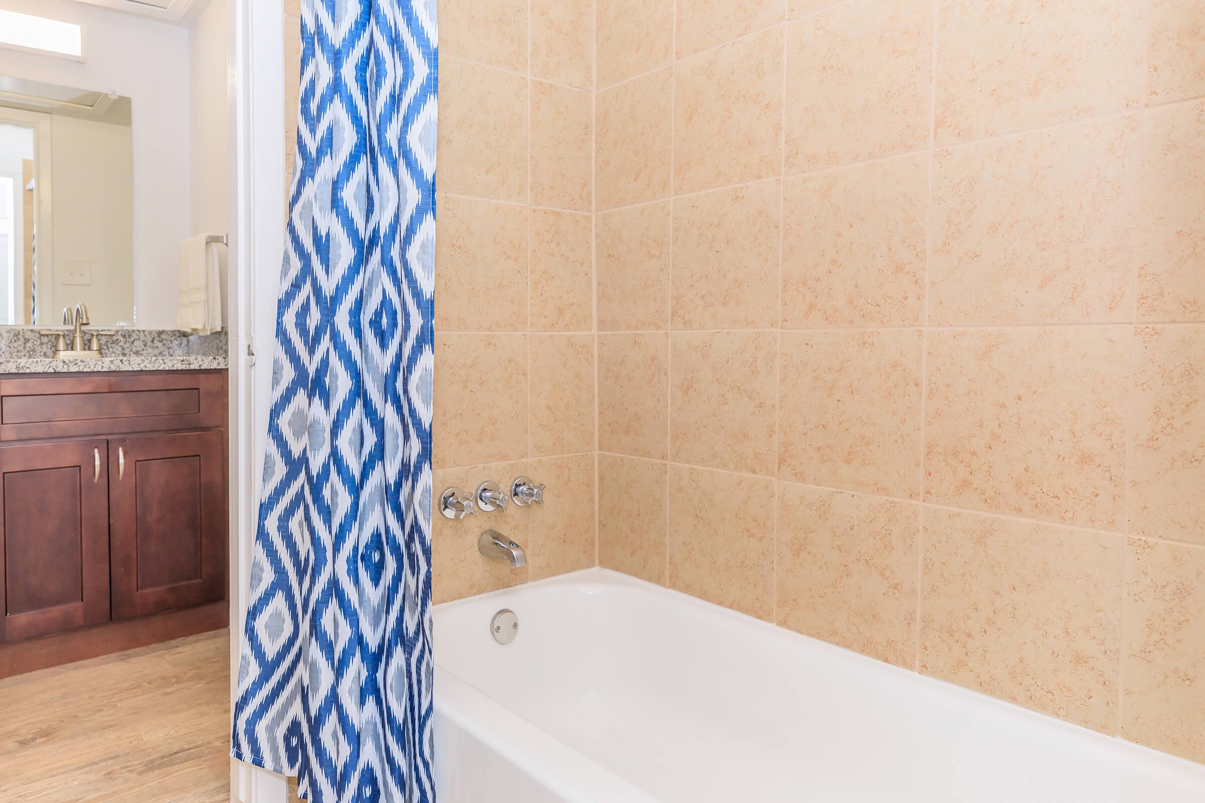 A bathroom scene featuring a white bathtub with a showerhead and faucet. A blue and white patterned shower curtain is partially drawn. The wall is tiled in beige, and there is a vanity with a mirror visible in the background. The flooring is wooden.
