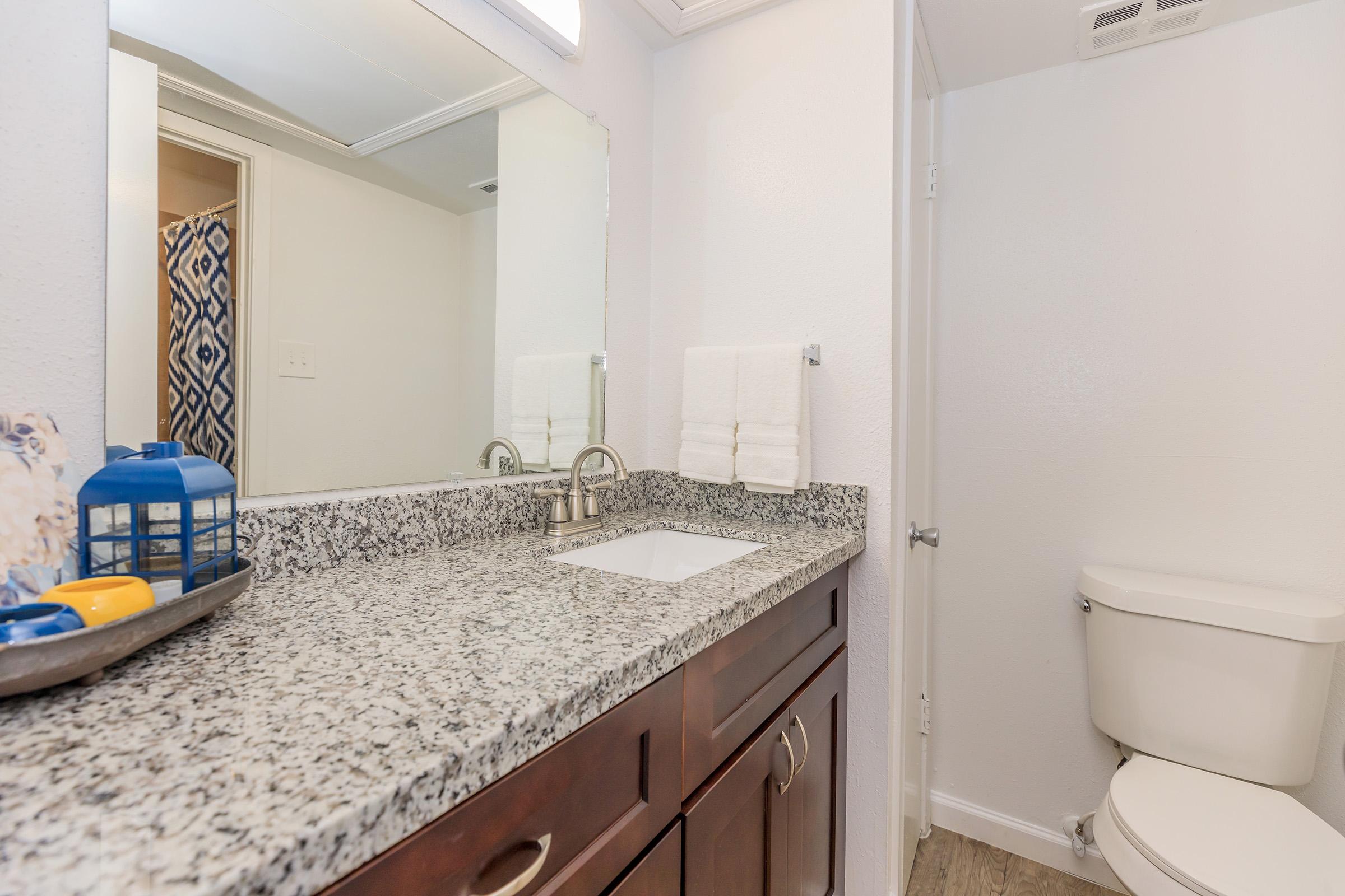 A clean bathroom featuring a granite countertop with a sink, two neatly folded white towels, and a decorative blue lantern. In the background, there is a shower curtain with a blue pattern, and a toilet is visible against the wall. The walls are painted a light color, creating a bright and airy atmosphere.