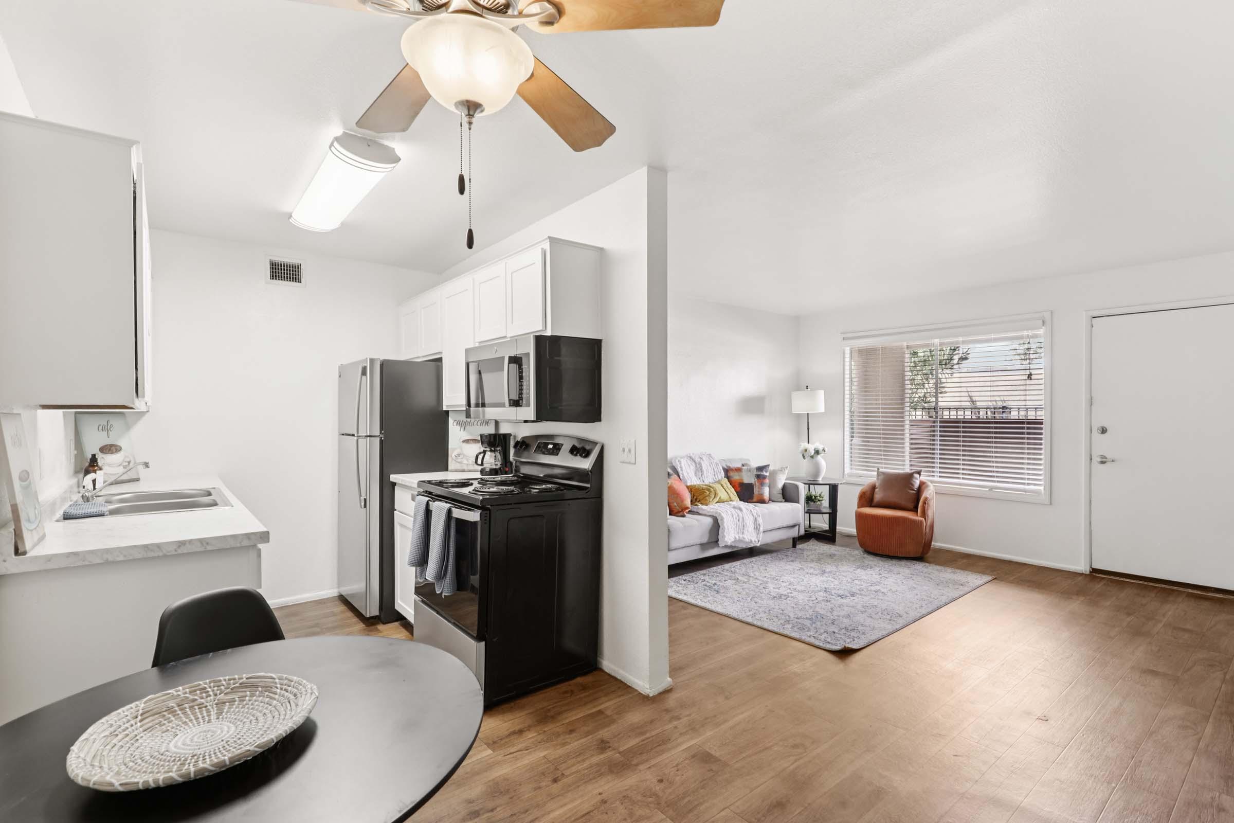 A modern apartment interior featuring a kitchen with white cabinets and stainless steel appliances, a dining area with a round table, and a cozy living room with a gray rug, orange and blue pillows on a sofa, and a brown armchair. Natural light enters through a window, enhancing the bright atmosphere.