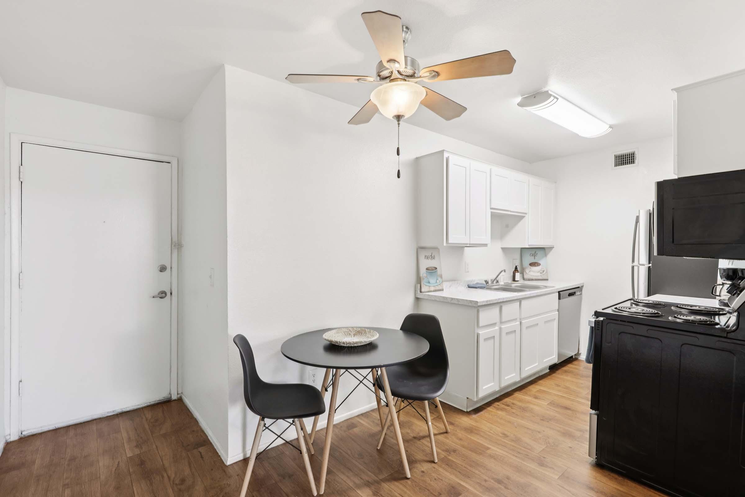 Modern kitchen featuring white cabinets, a small round table with black chairs, and stainless steel appliances. A ceiling fan is visible above, and there is a doorway leading to another room. The floor is a light wood finish, creating a bright and inviting atmosphere.