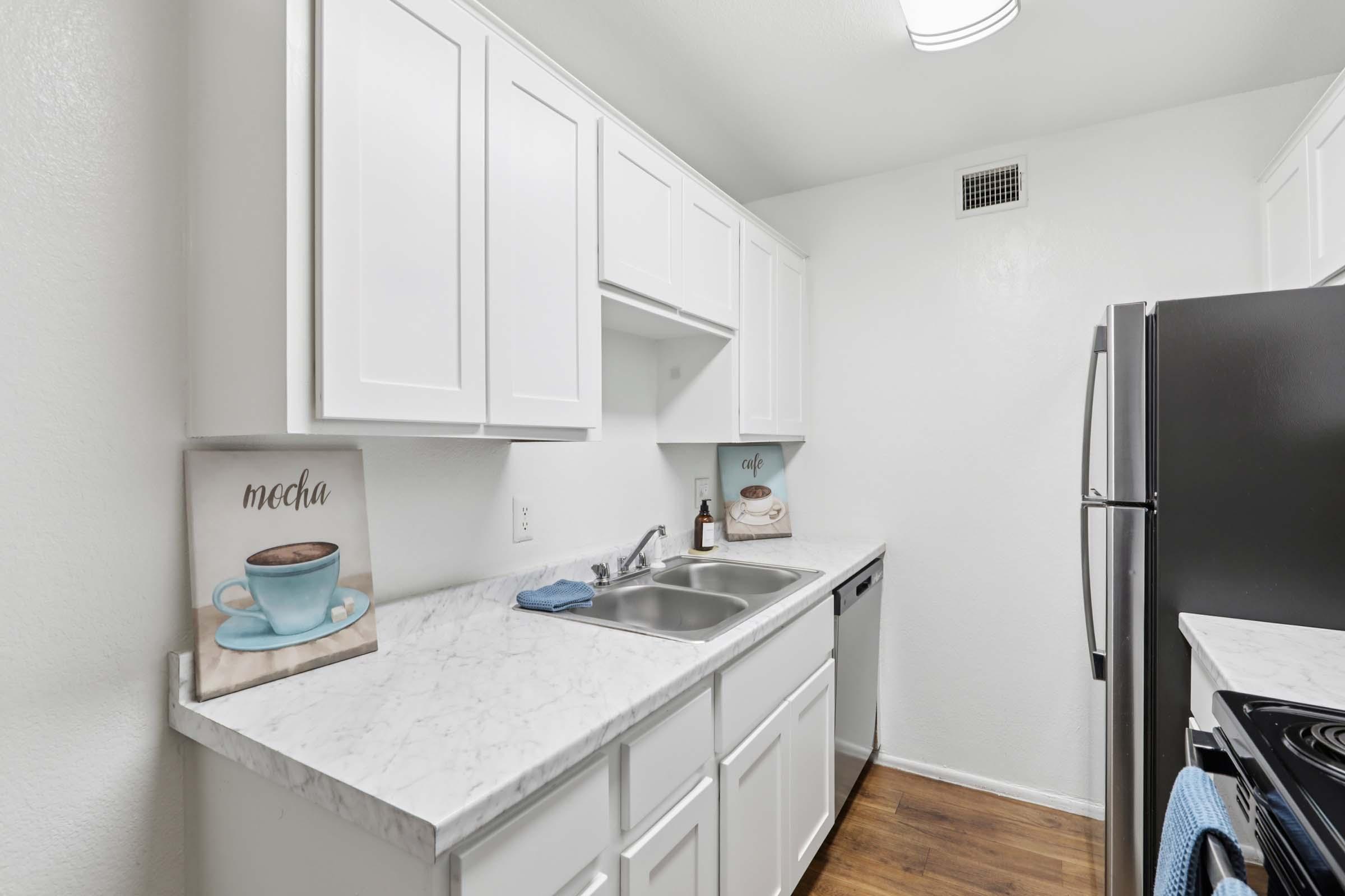 Modern kitchen with white cabinetry, a marble countertop, and stainless steel appliances. Featuring a double sink and a black stove. Two decorative signs on the counter read "mocha" and "café." The walls are painted white, and there is a window providing natural light.