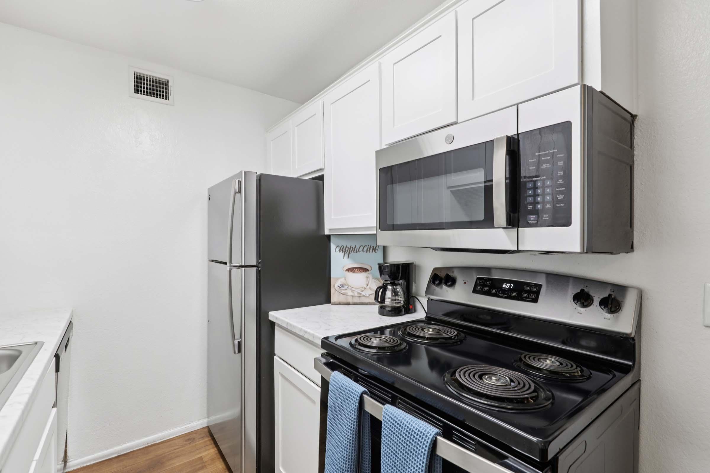 A modern kitchen featuring white cabinets, a stainless steel refrigerator, a black stove with an oven, and a microwave above. The countertops are light-colored, and there's a coffee maker on the counter. The flooring is wooden, contributing to a contemporary look.