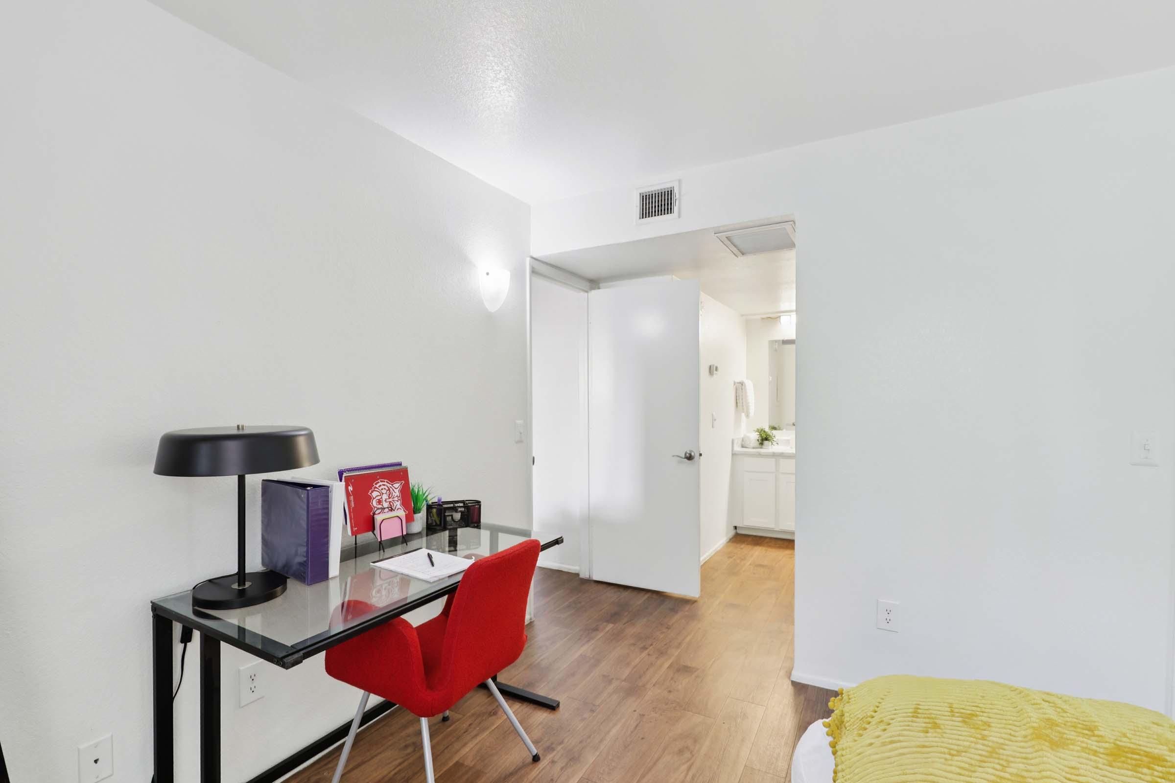 A bright, minimalist room featuring a small desk with a black lamp, a red chair, and some colorful folders. There is a white door leading to a bathroom, light wood flooring, and a bed with a yellow throw blanket, creating a cozy and inviting atmosphere.