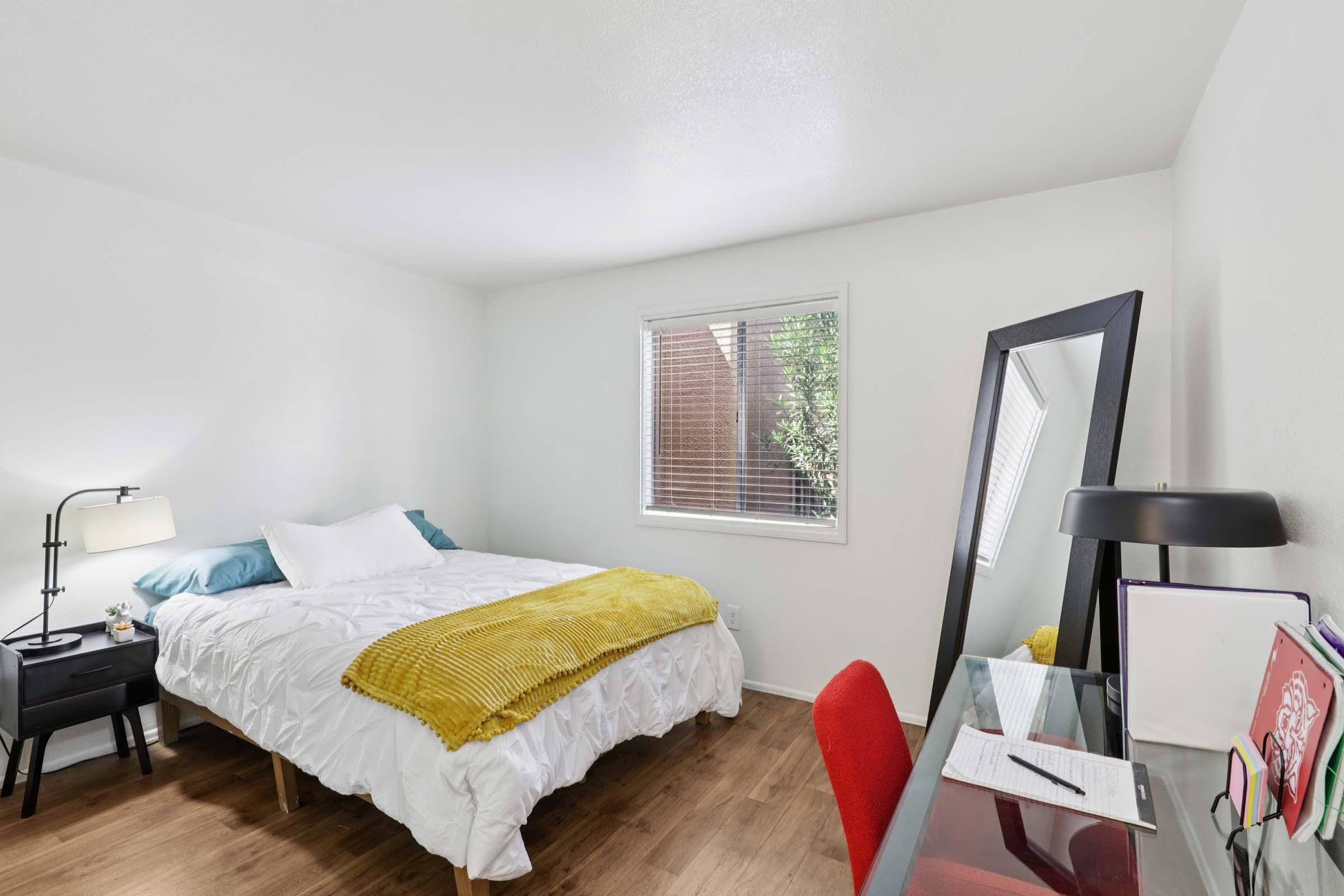 A simple and bright bedroom featuring a double bed with white linens and a yellow throw blanket. There's a small nightstand with a lamp, a large mirror, and a desk with a red chair. A window with blinds allows natural light to fill the space, complemented by wooden flooring.