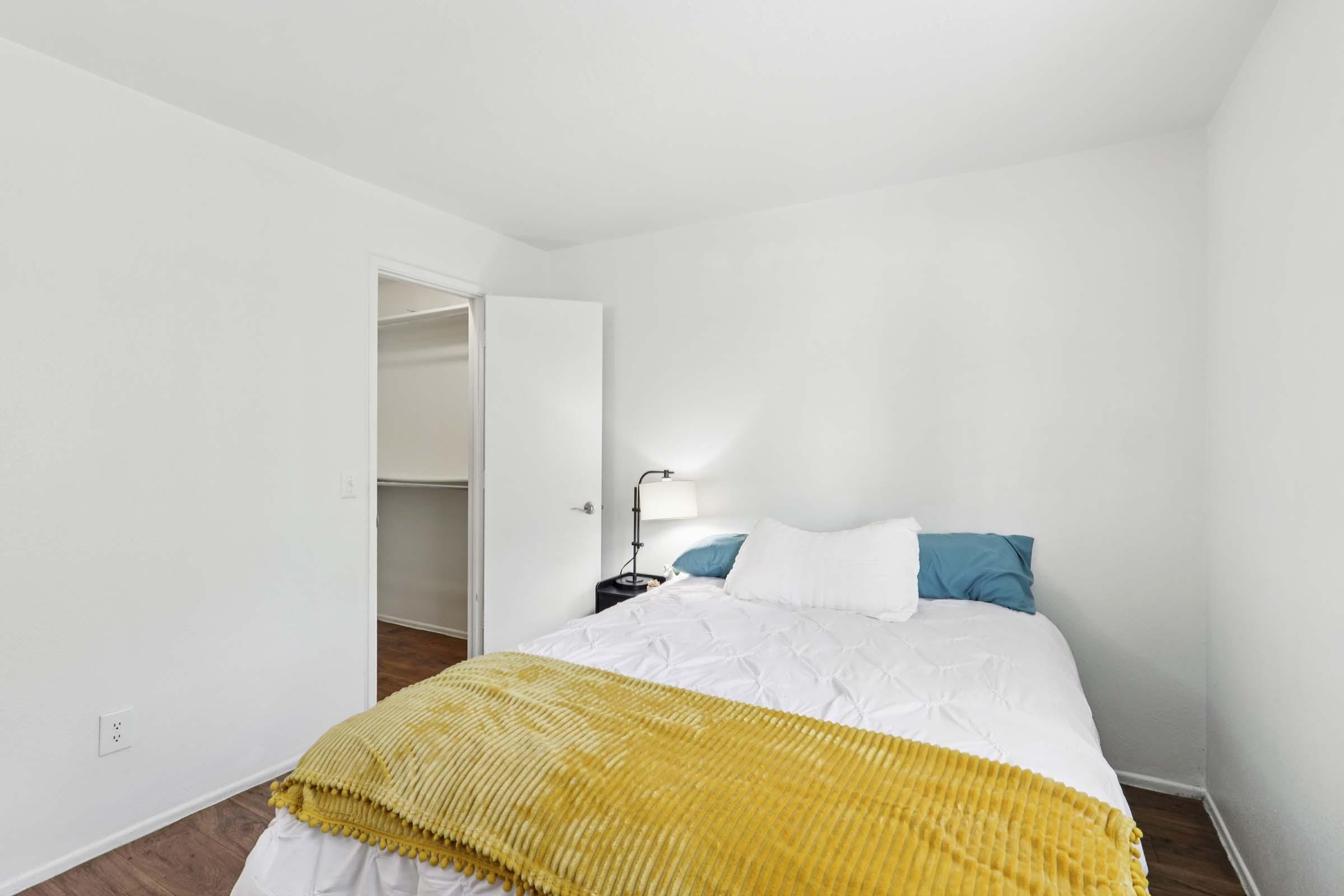 A cozy bedroom featuring a white bed with blue and white pillows, a yellow throw blanket, a bedside lamp, and a closet with an open door. The walls are white, and the flooring is wooden. The room is well-lit and minimalistic, creating a calm atmosphere.