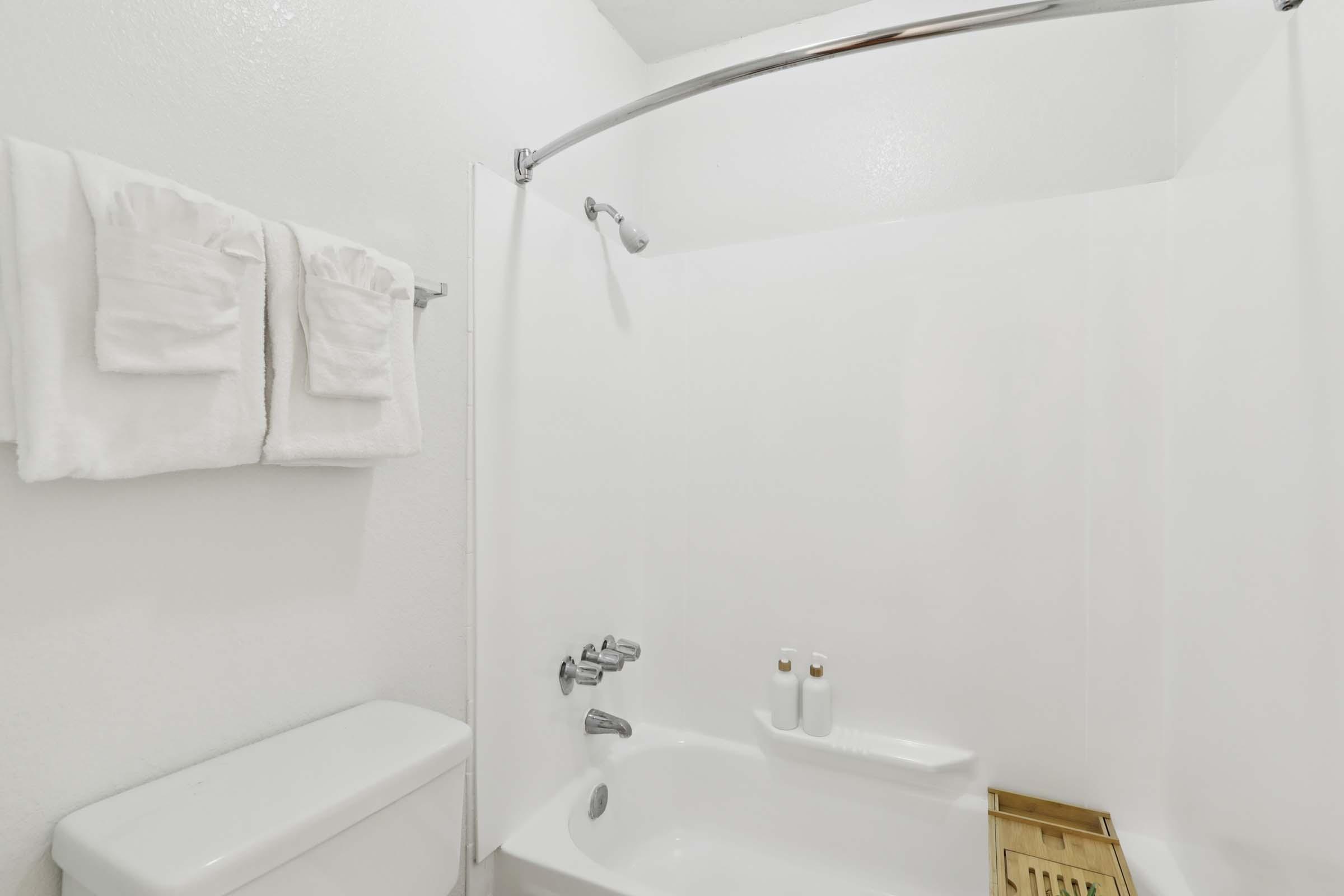 A clean and simple bathroom with a shower and bathtub combo. The walls are white, and there are two folded white towels hanging on a rod. A showerhead is mounted above, and a small shelf with two bottles is visible on the tub's edge. A gold-colored bath mat is placed on the floor.