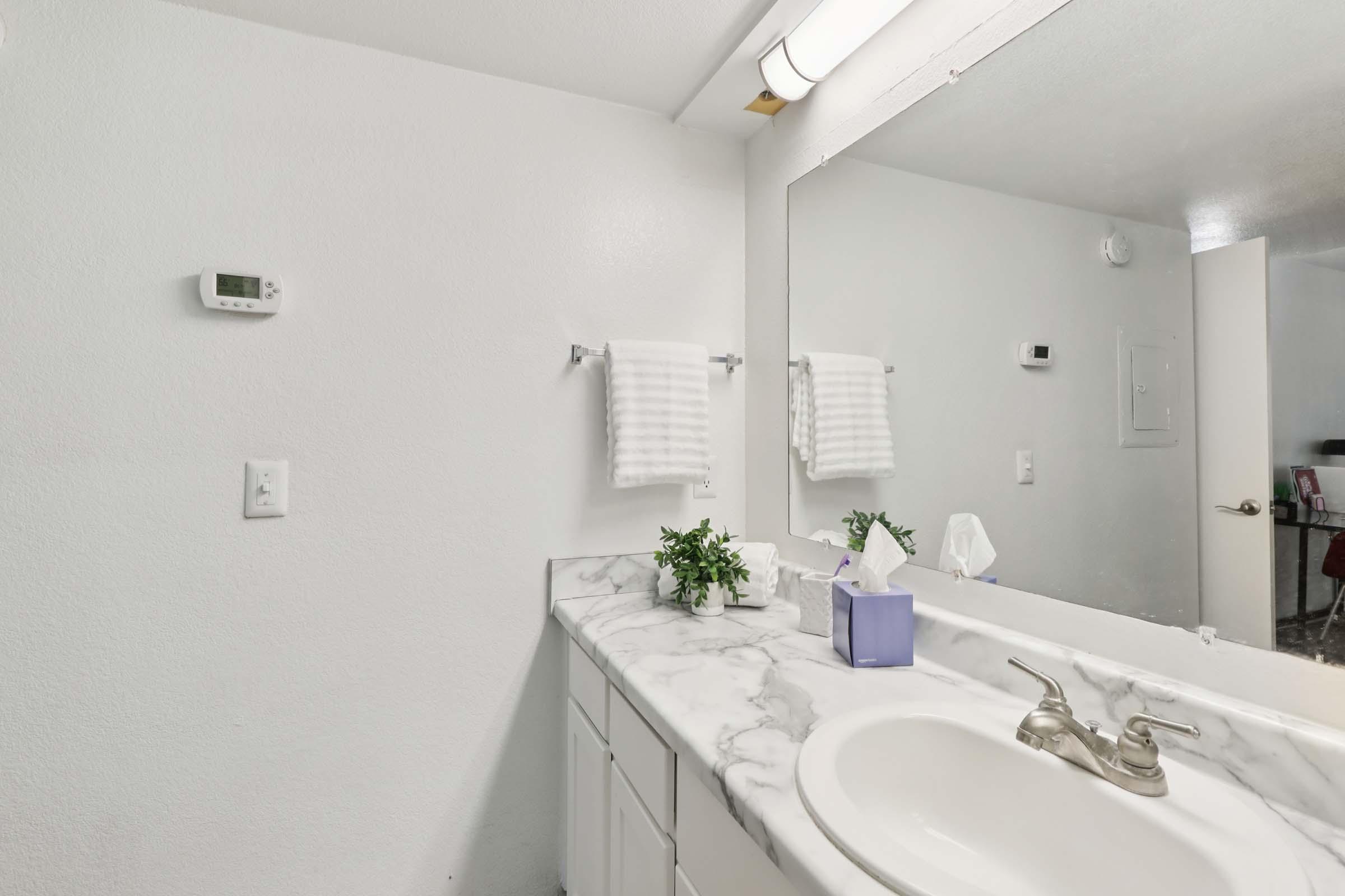 A clean and modern bathroom featuring a white marble countertop with a sink, a large mirror, and neatly hung white towels. In the corner, there are small green plants, and a tissue box is placed next to the sink. The wall is painted white, and there are visible light fixtures and a thermostat.