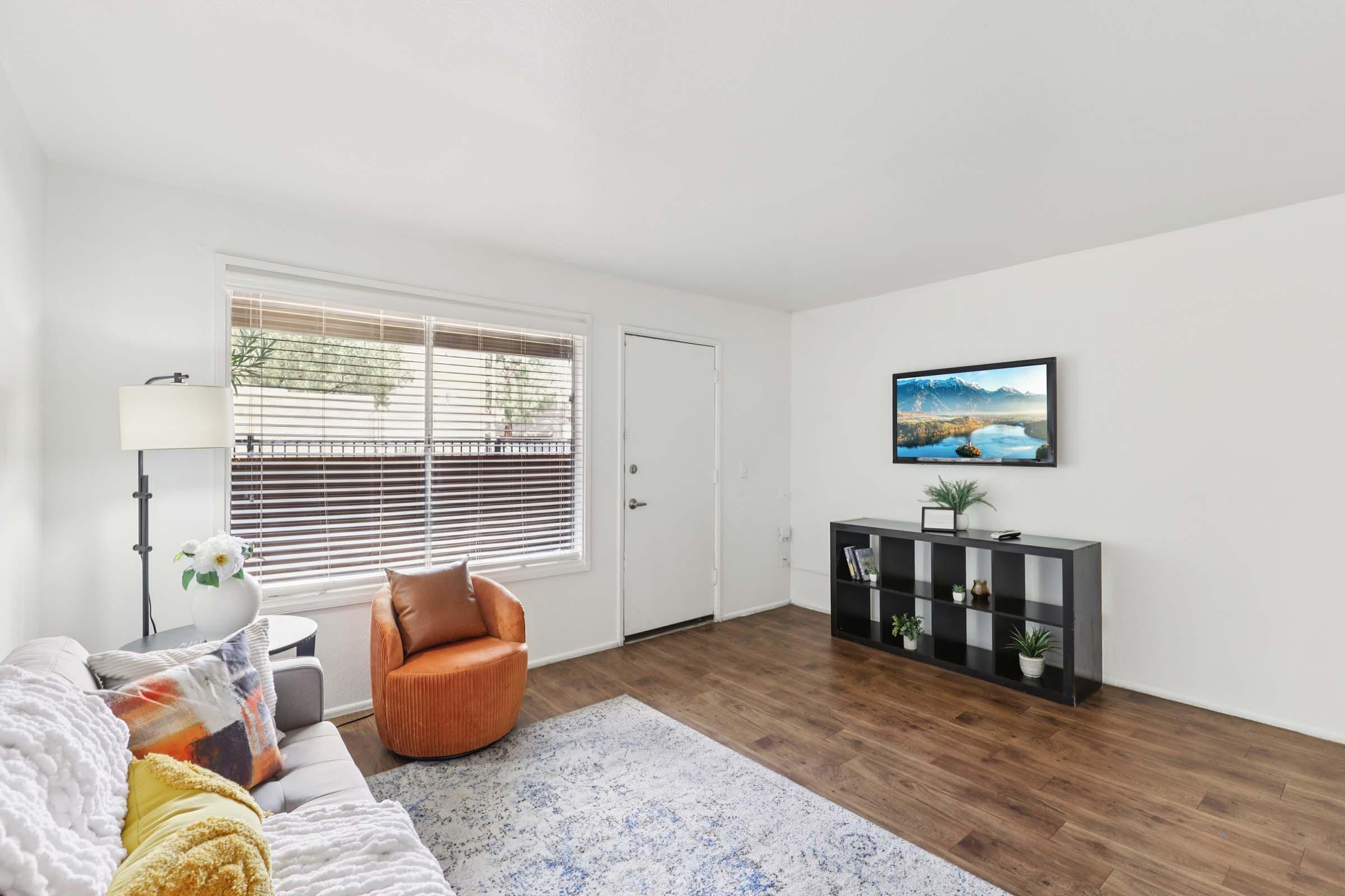 Bright and modern living room featuring a beige sofa with colorful throw pillows, an orange armchair, a black TV stand with decorative plants, and a television displaying a serene landscape. Large window with blinds allows natural light to fill the space, complemented by warm wooden flooring.