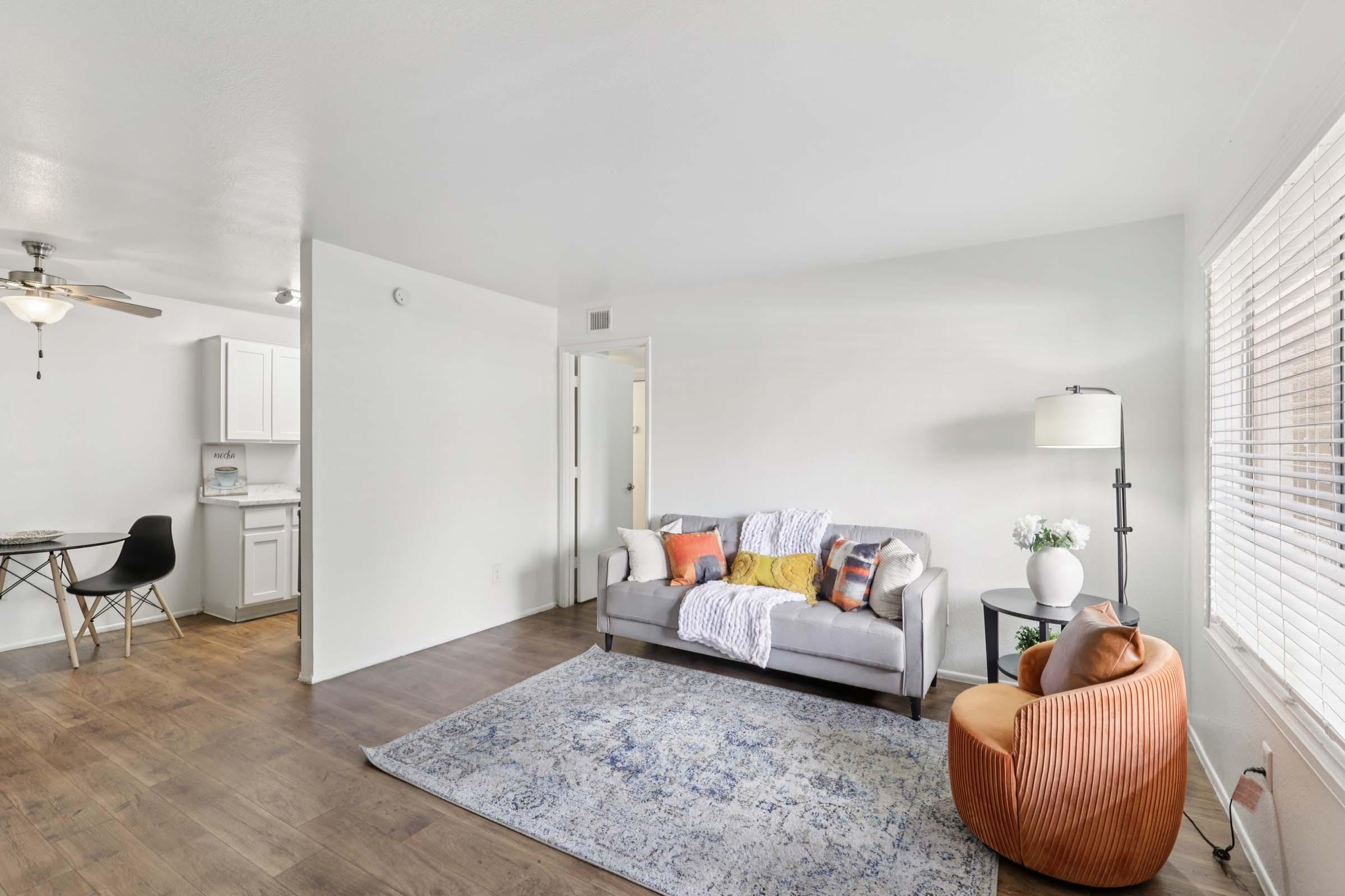 A cozy living room featuring a light gray sofa adorned with colorful throw pillows, a decorative orange chair, and a patterned area rug. A small dining table with black chairs is visible in the background, along with a well-lit kitchen area. The walls are painted white, creating a bright and inviting atmosphere.