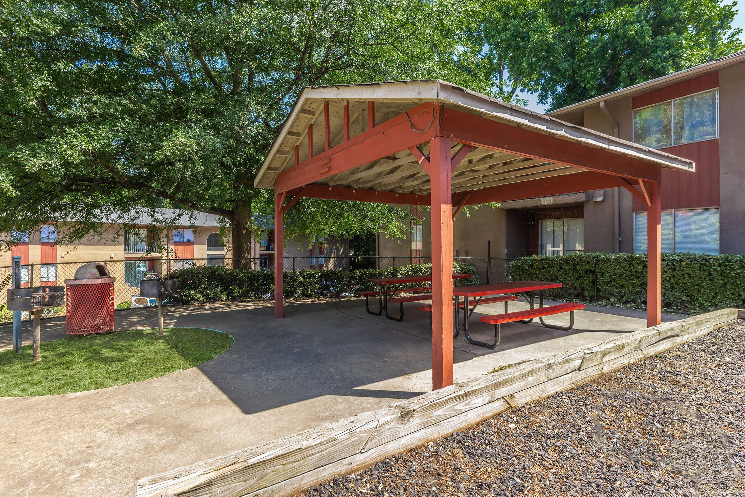 A shaded outdoor pavilion with a red wooden frame and picnic tables underneath, surrounded by green trees and landscaped grass. In the background, a fenced area and a multi-story building are visible. The setting is peaceful and inviting for gatherings.