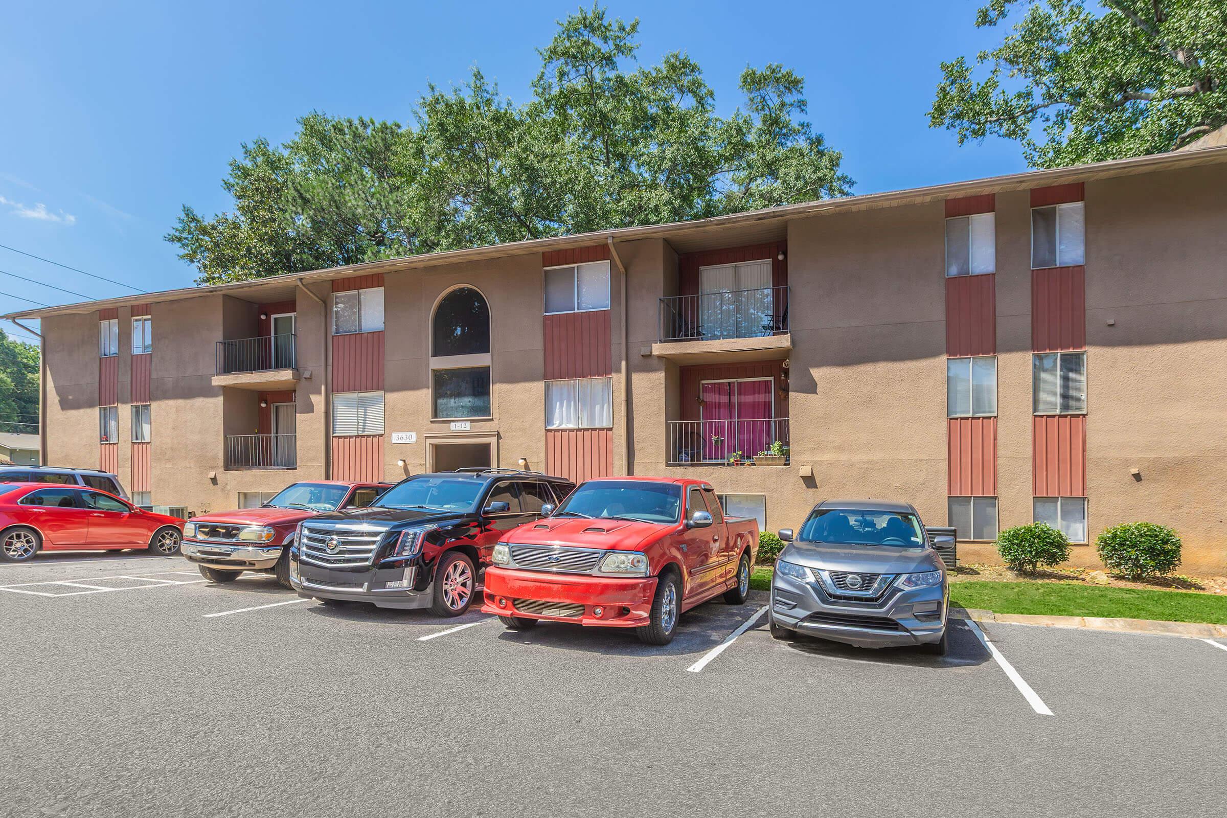 View of a two-story apartment building with a beige exterior and red accents. Several parked cars in the foreground, including a red truck, a dark SUV, and a silver sedan. Green trees and bushes frame the scene under a clear blue sky.