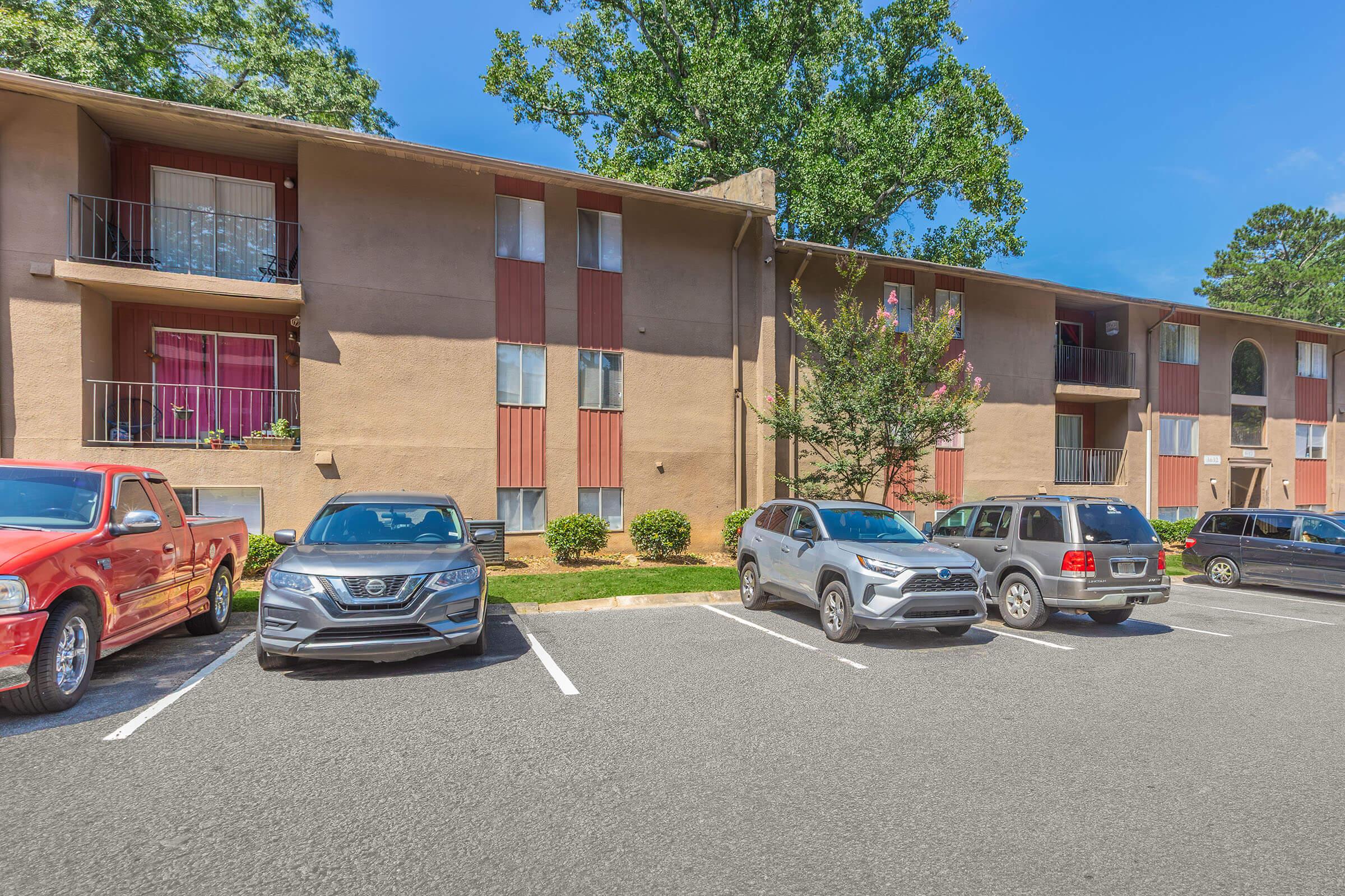 Five parked cars in front of a two-story apartment building with a tan exterior and red accents. The building has balconies with some greenery around it, under a clear blue sky. There are trees in the background, creating a pleasant residential atmosphere.