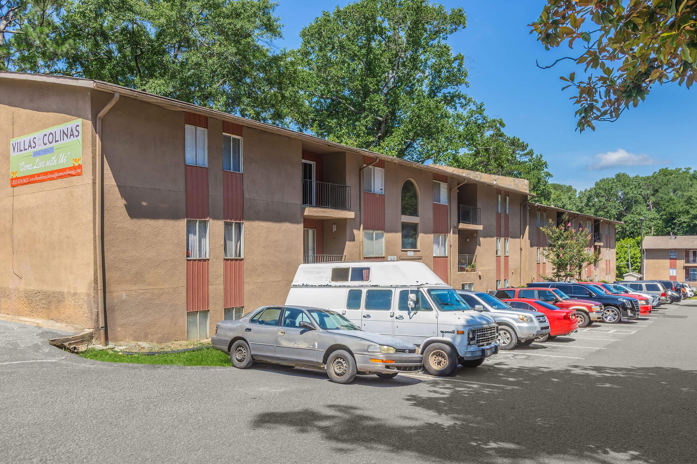 Exterior view of a multi-story apartment building named "Villas Colinas," with a sign visible. A row of parked cars, including a white van and various sedans, line the parking area in front of the building. The scene is set against a backdrop of green trees and a clear blue sky.