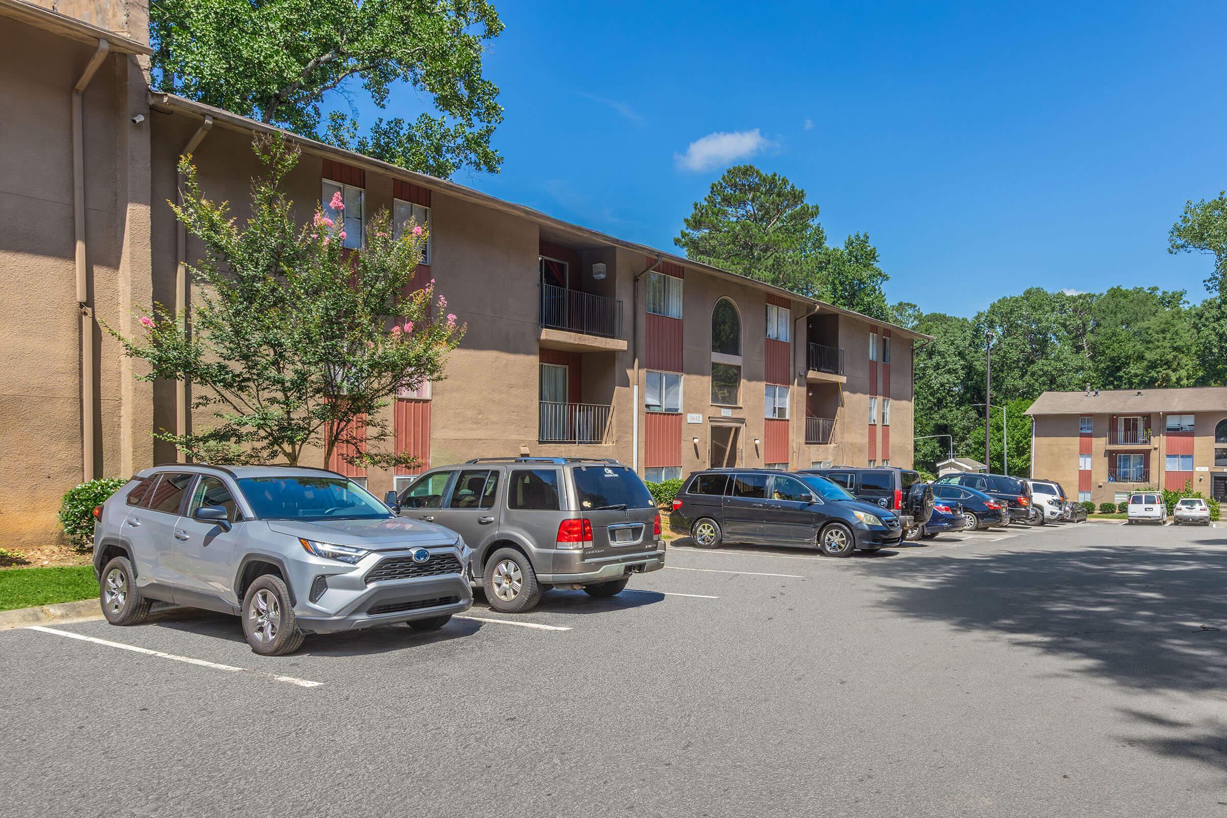 A view of an apartment complex with two buildings. The foreground shows a parking lot with several cars, including a silver SUV and a gray sedan. Green trees and a clear blue sky are visible in the background, contributing to a sunny, suburban atmosphere.