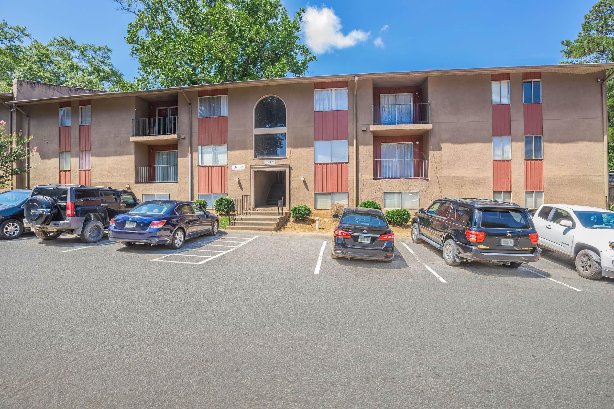 A two-story apartment building with a tan exterior, featuring red accents on the balconies. Several parked cars are visible in the foreground, including black SUVs and a gray vehicle. The setting includes a grassy area and trees, under a clear blue sky with a few clouds.