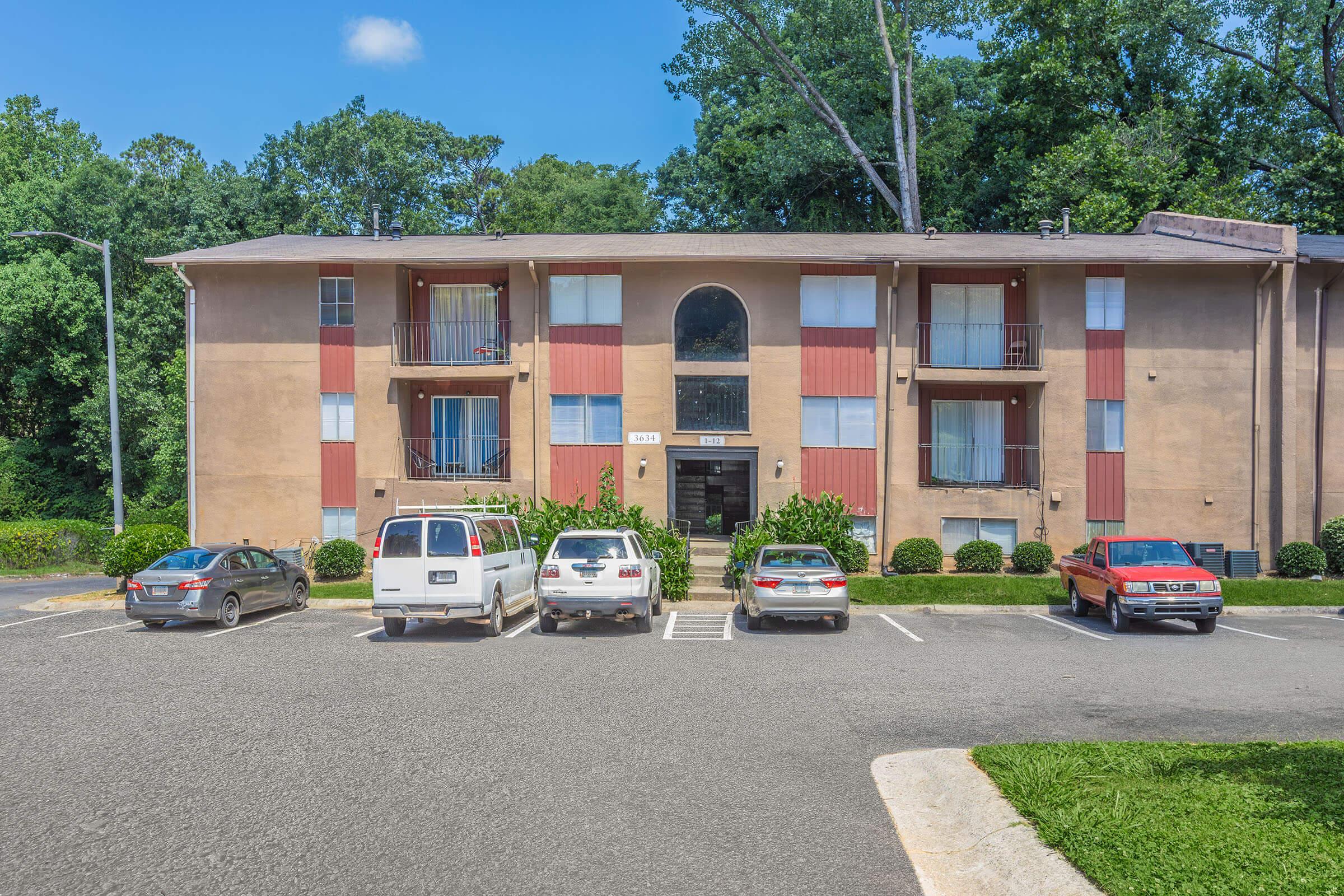 A three-story apartment building with a tan exterior and red accents, surrounded by trees. The front features a large arch window and several balconies. In the parking lot, there are several parked vehicles, including a white van and multiple sedans. Clear blue sky overhead.