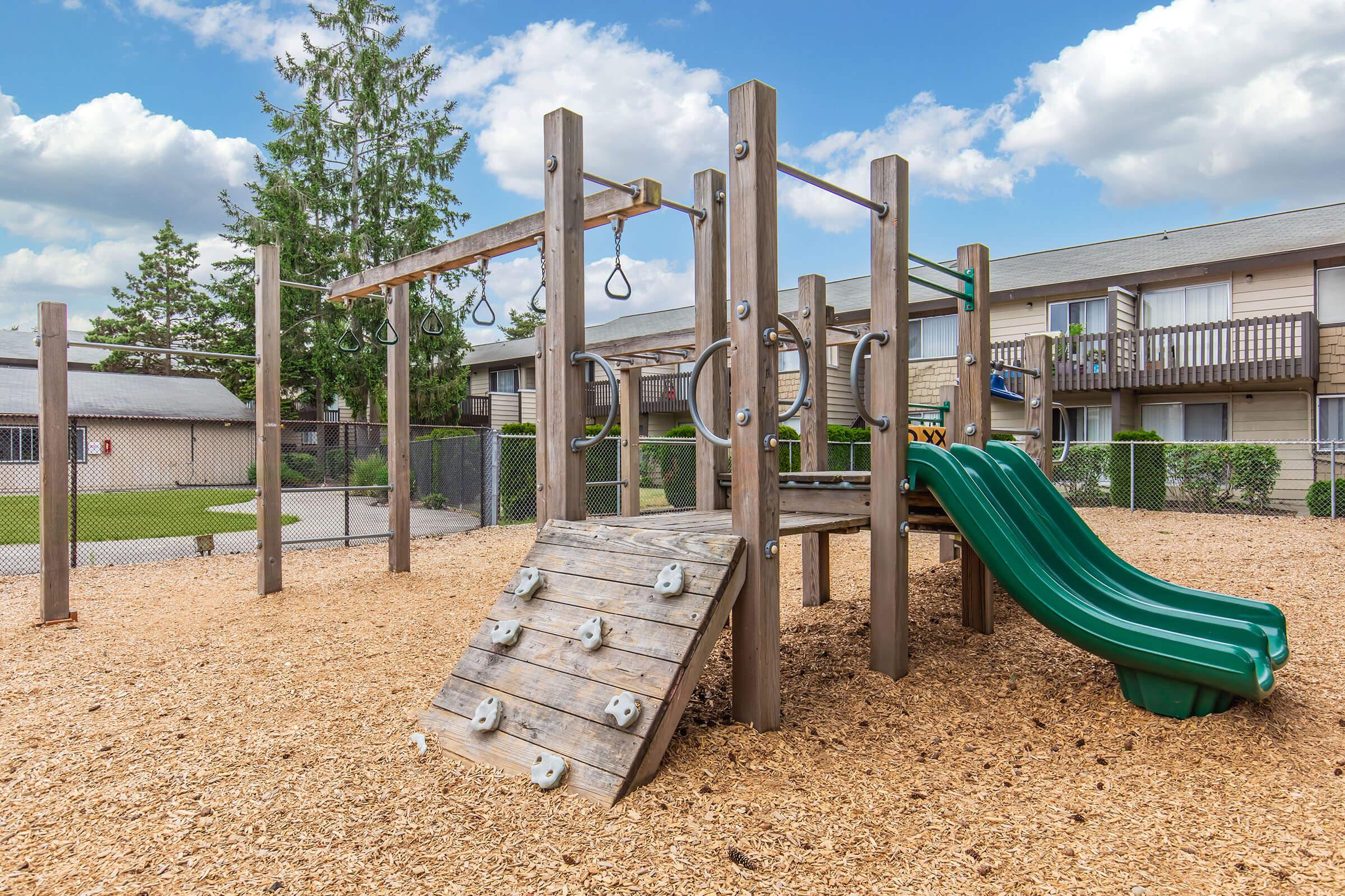 A playground structure featuring a climbing wall, swings, and a green slide, surrounded by wood chips. In the background, there are several apartment buildings with balconies and trees under a partly cloudy sky.