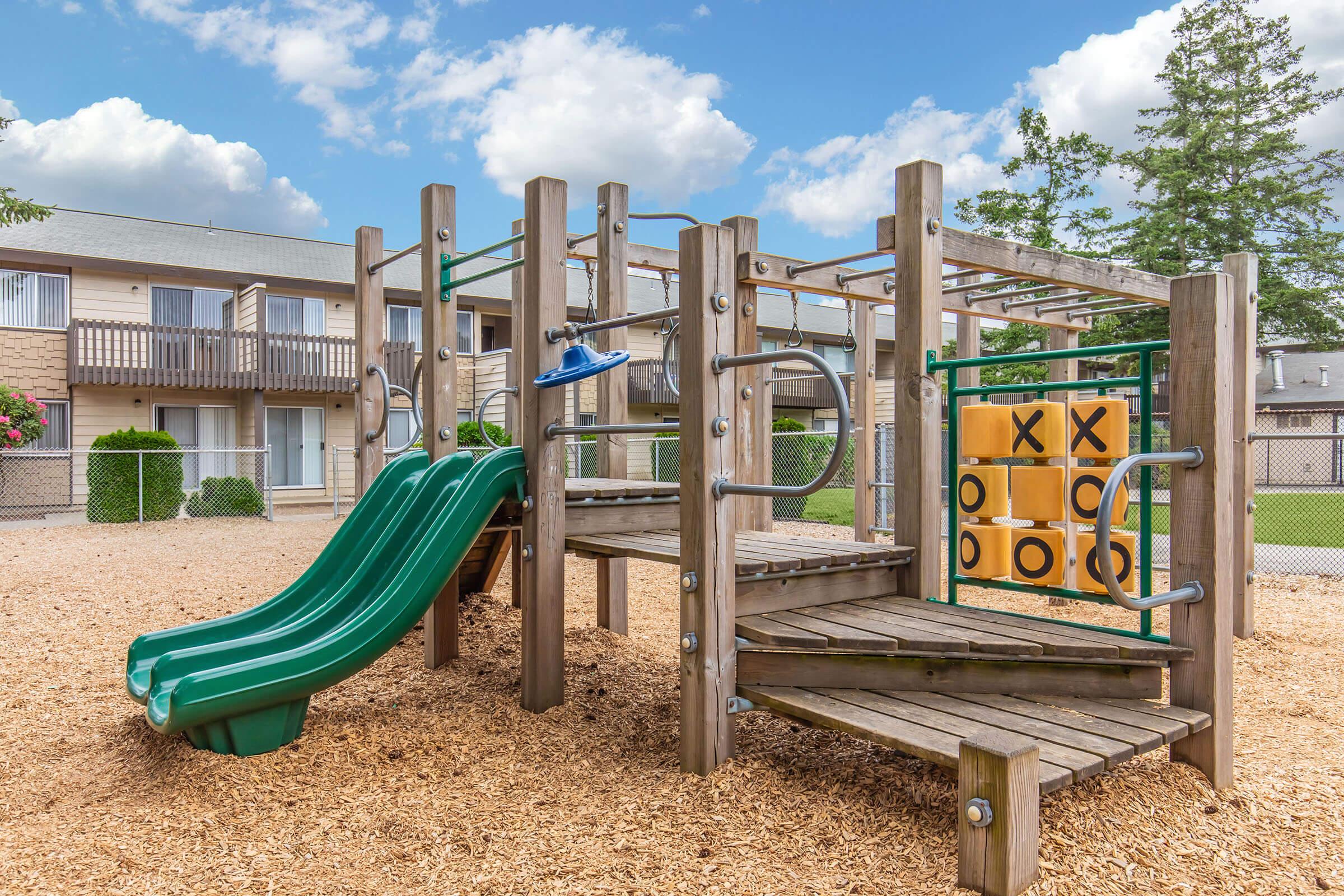 A children’s playground featuring a green slide, climbing structures, and a tic-tac-toe board. The area is surrounded by wood chips and is set against a backdrop of an apartment building with balconies. The sky is partly cloudy, adding a bright atmosphere to the scene.
