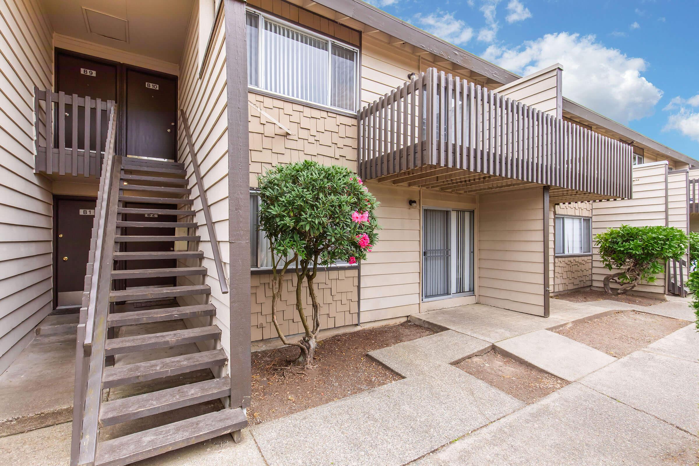 Exterior view of a multi-unit apartment building featuring two stories. A wooden staircase leads to the upper level, which has a balcony. The building has a light-colored exterior with textured wall panels and several windows. There are small shrubs and landscaping in front of the entrance.