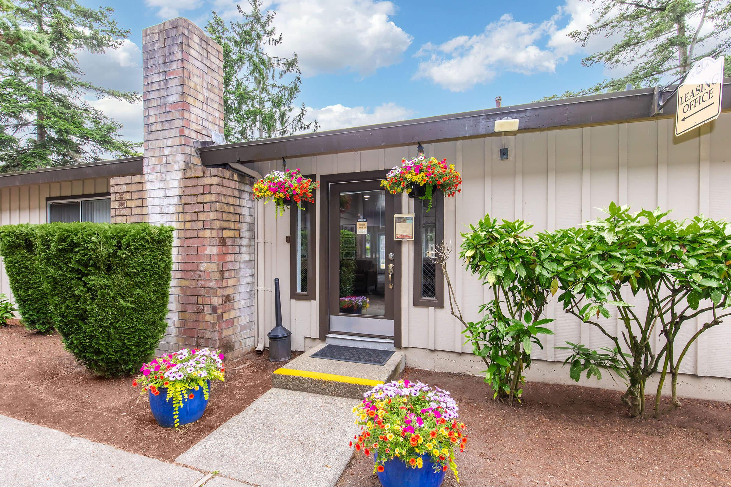 Exterior view of a rental office with a welcoming entrance. The building features a brick chimney and is surrounded by lush greenery and colorful flower pots. Flower baskets hang near the entrance, adding a vibrant touch to the overall appearance. A clear blue sky is visible in the background.