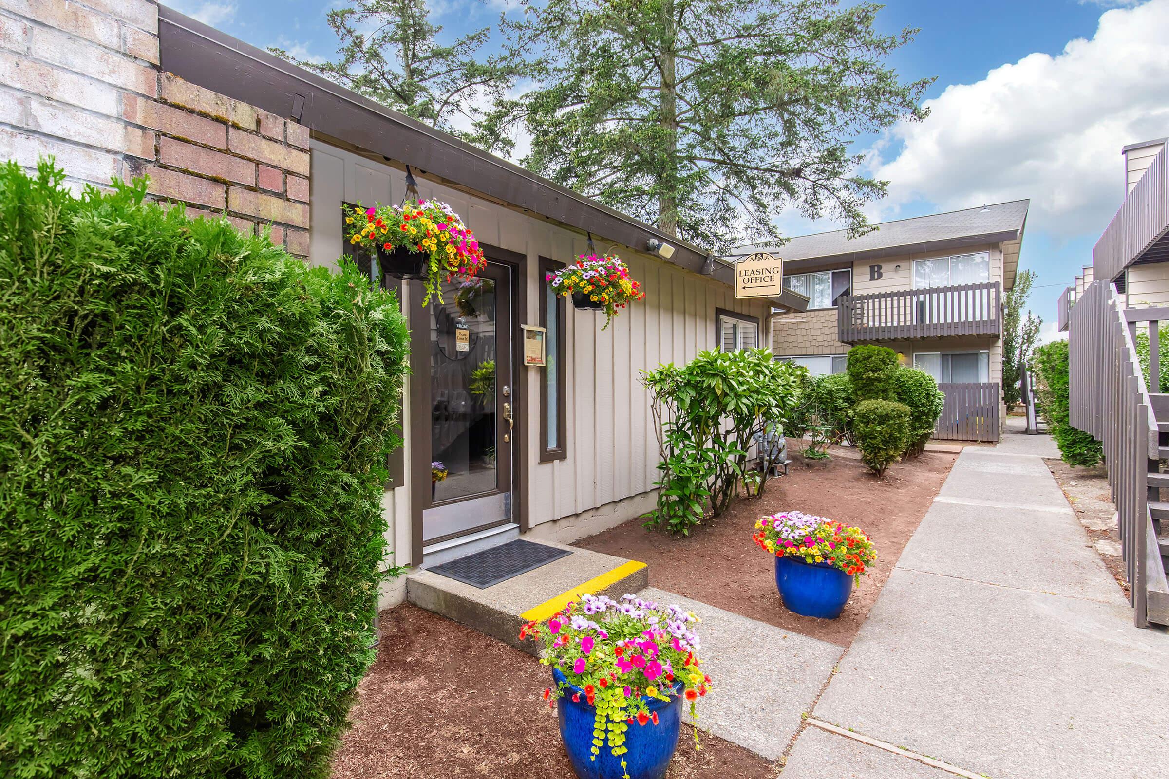 A welcoming entrance to a building, featuring a door with a business sign, surrounded by neatly trimmed hedges and colorful flower pots in blue planters. The pathway leads to different units, with a glimpse of greenery and a blue sky above.