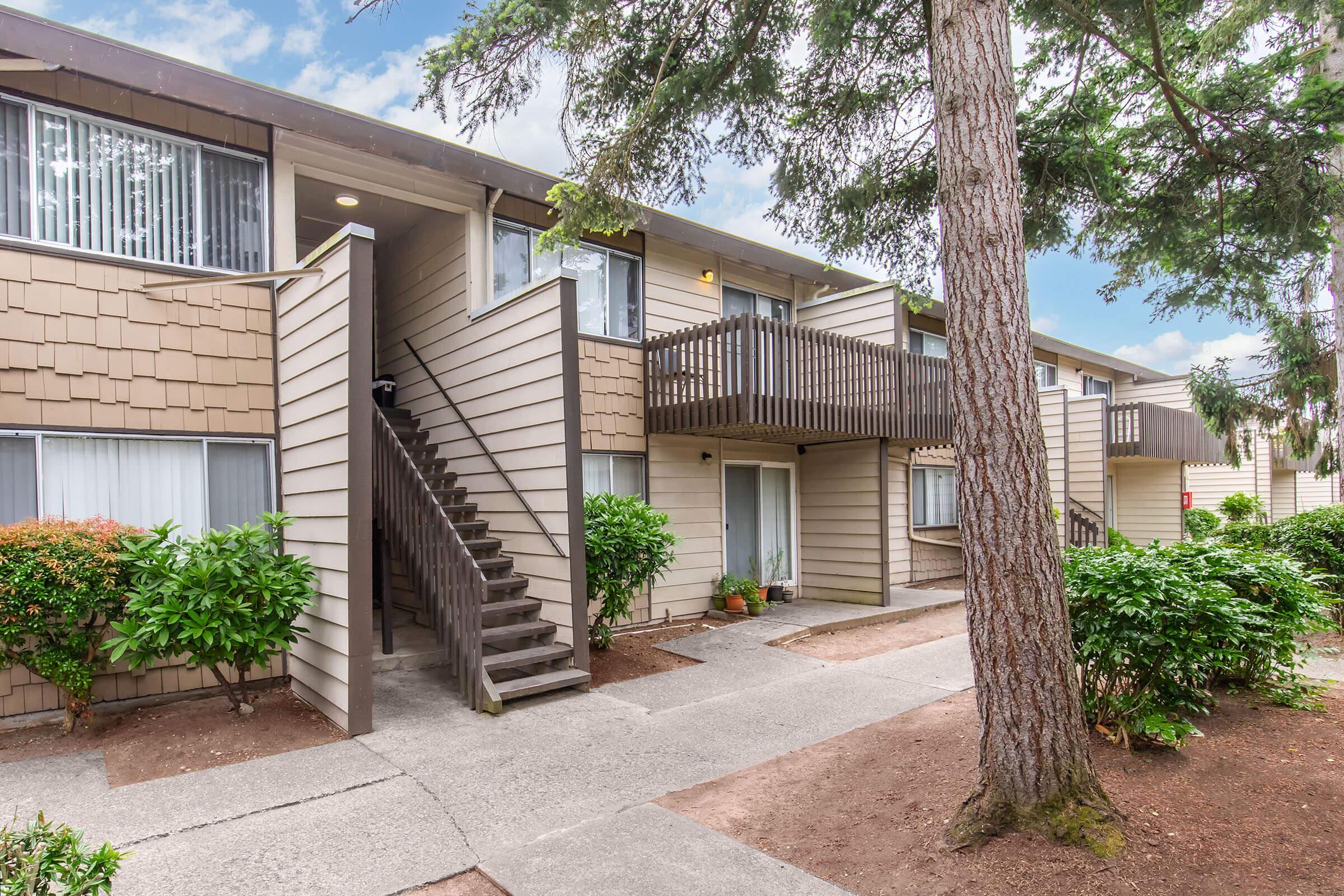 A multi-unit residential building with a light brown exterior, featuring a staircase leading to an upper level. The landscape includes shrubs and trees, and there's a clear blue sky above. The walkway is paved, leading to the entrance of the building.