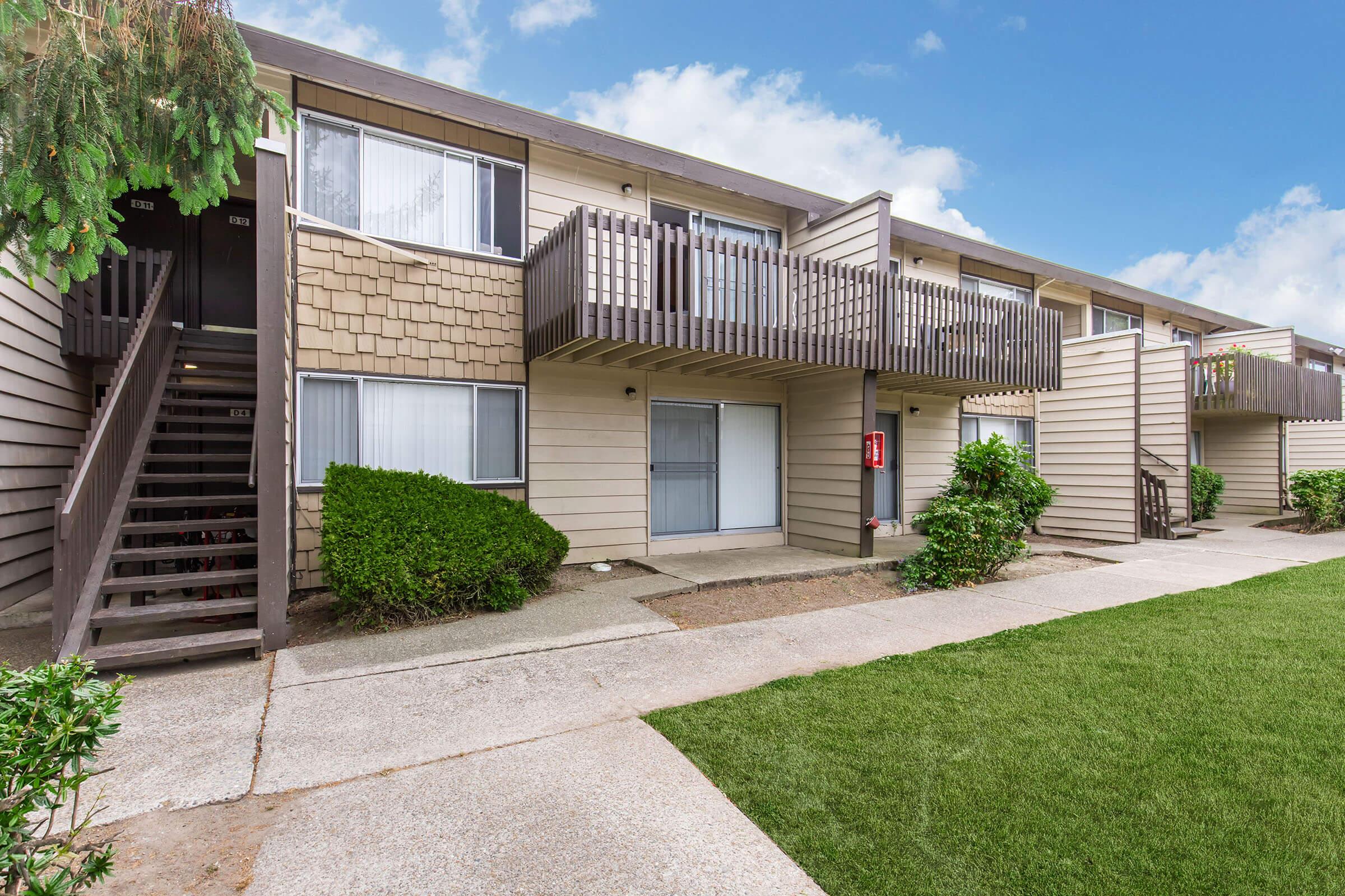 A row of two-story apartment buildings with beige siding and wooden balconies. Grass-lined pathway leads to the entrance. Nearby are small bushes and a set of stairs leading to the upper level. The sky is partly cloudy, giving a bright, welcoming ambiance.