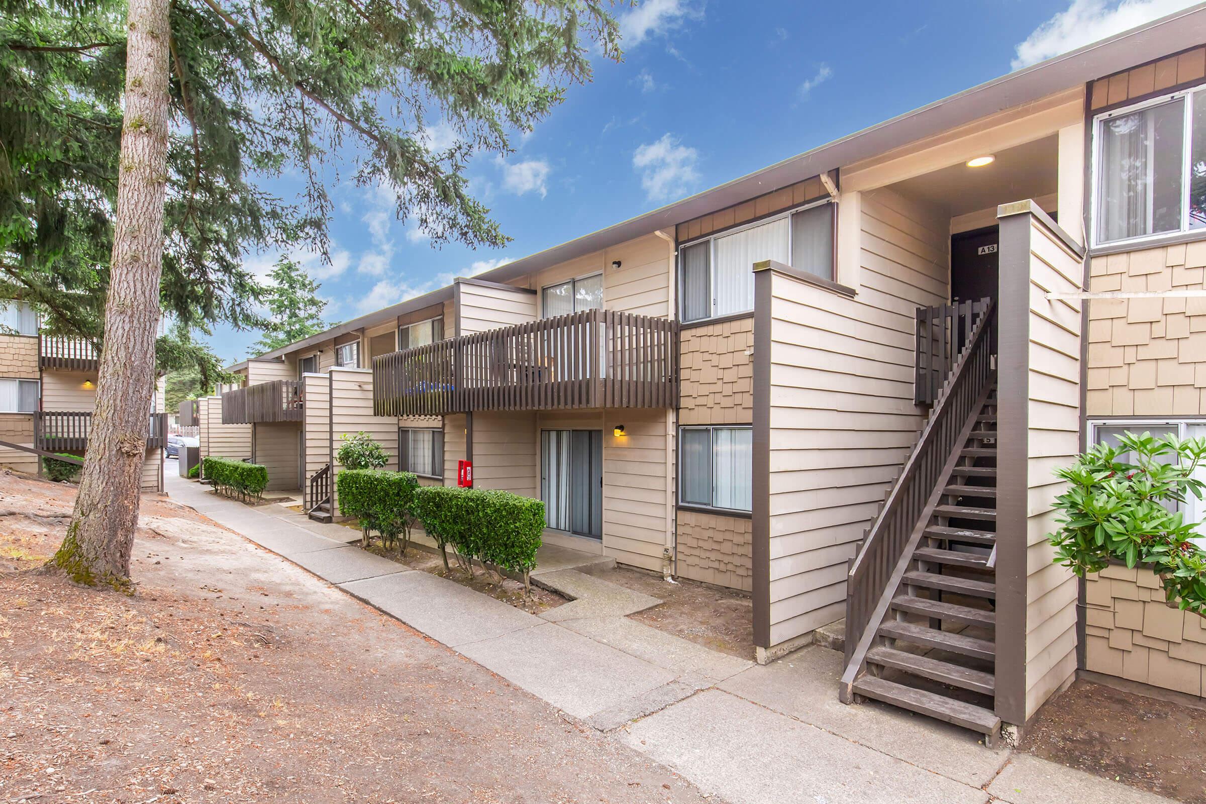 A multi-unit residential building with a light beige exterior and wood accents. The two-story structure features balconies with railings, a staircase leading to upper units, and landscaped greenery along the sidewalk. The sky is blue with some clouds, creating a pleasant atmosphere.