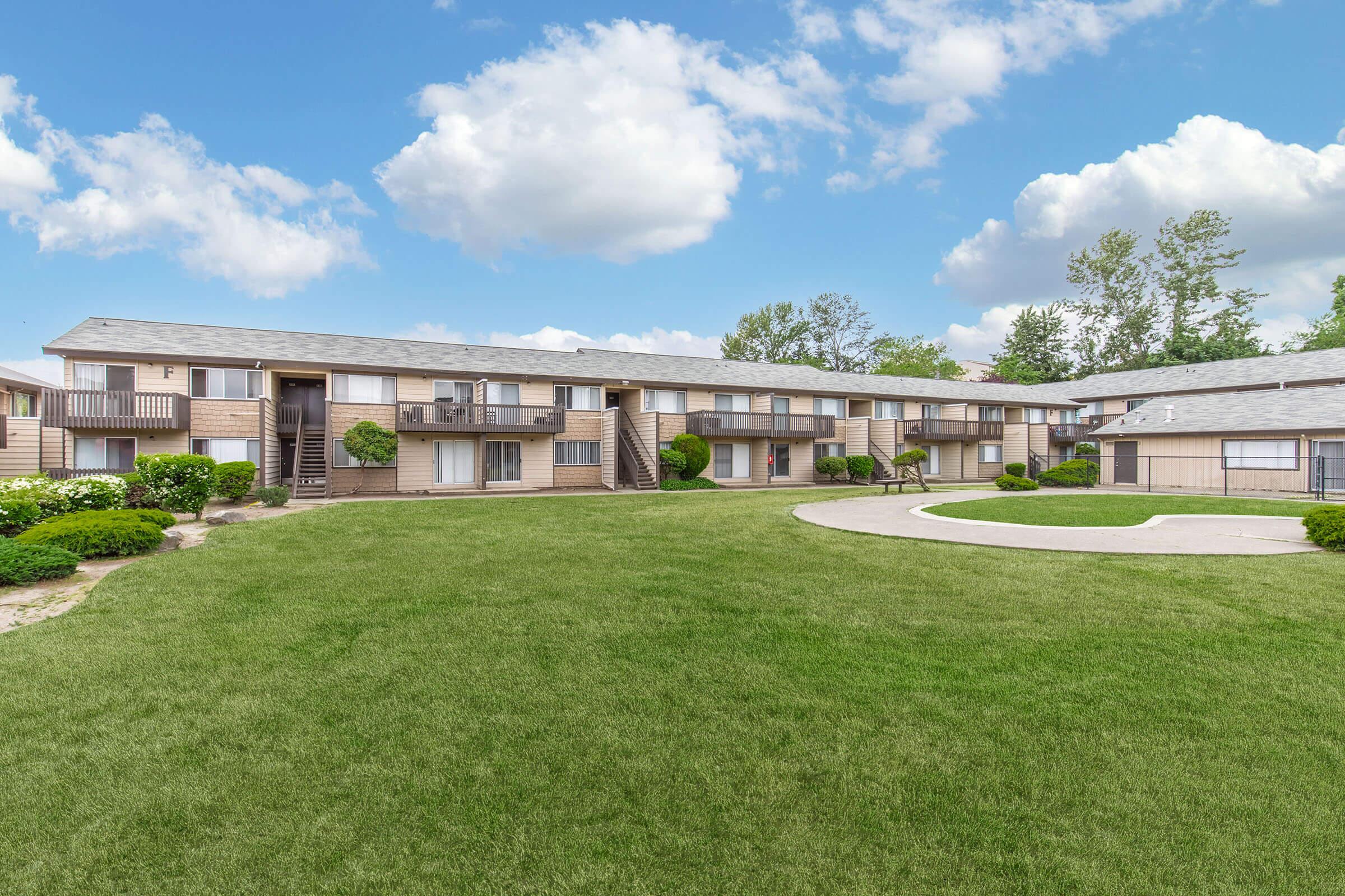 A well-maintained apartment complex with two-story buildings surrounding a green lawn. The landscape features trimmed bushes and a circular pathway. The sky is bright with fluffy clouds, creating a welcoming atmosphere.