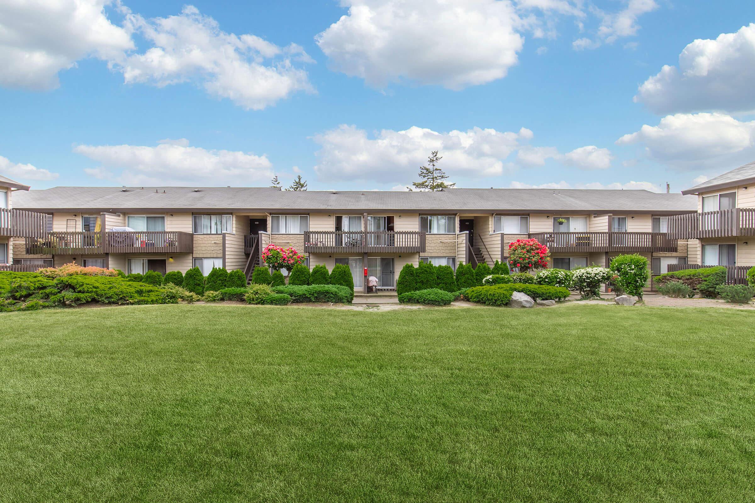 A view of an apartment complex featuring two-story buildings with balconies. Lush green grass covers the foreground, bordered by manicured shrubs and flower beds with vibrant blooms. The sky is partly cloudy, creating a bright and inviting atmosphere.