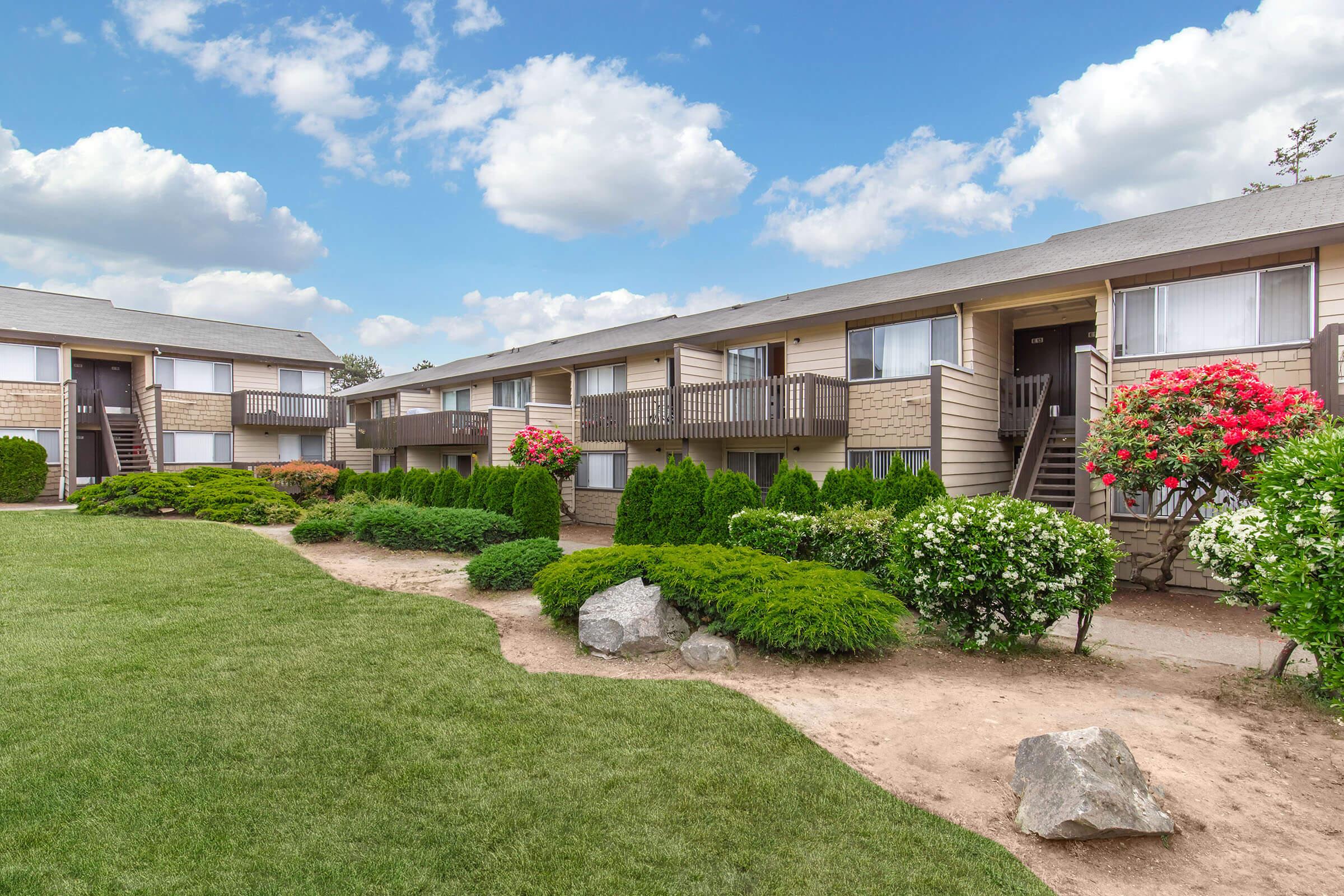 A row of two-story apartment buildings with balconies, surrounded by well-maintained grassy areas and colorful flower bushes. The sky is partly cloudy, adding a bright atmosphere to the scene. Pathways lead up to the entrances of the apartments, providing a welcoming feel.