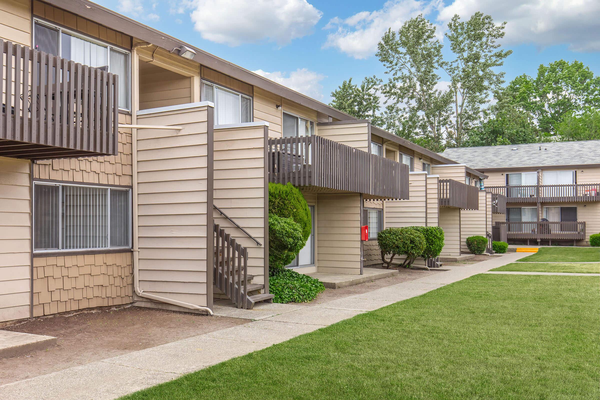 A view of a residential apartment complex featuring two-story buildings with balconies, well-maintained lawns, and neatly trimmed bushes. The sky is partly cloudy, adding a pleasant backdrop to the scene. Walking paths lead through the grassy area, contributing to a serene living environment.