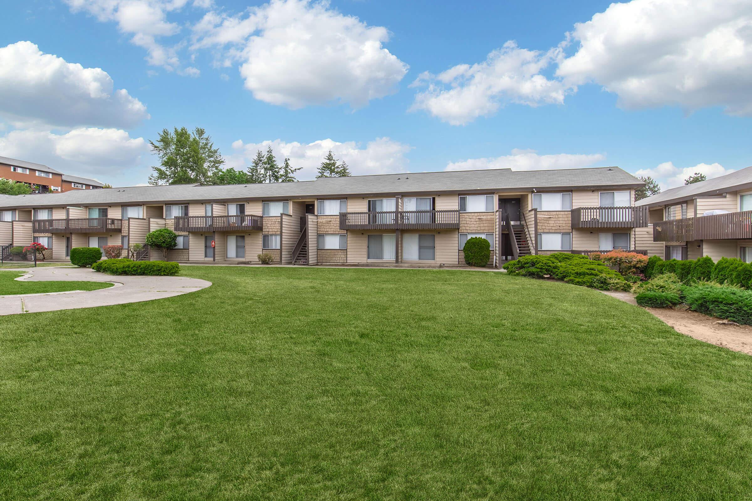 A landscaped courtyard featuring a two-story apartment building. The building has multiple balcony units and is surrounded by well-maintained grass and shrubs. The sky is partly cloudy, creating a pleasant atmosphere.