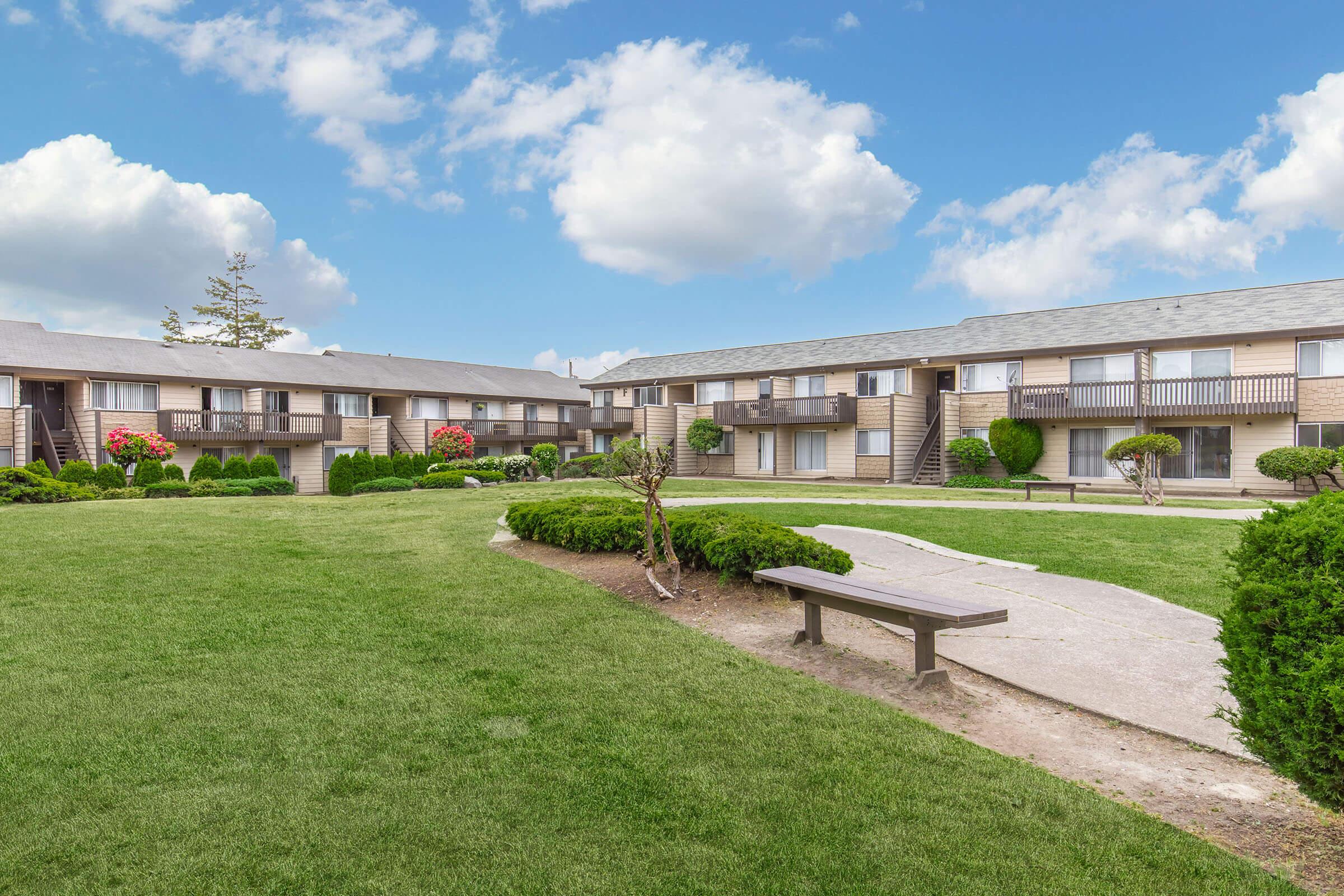 A well-maintained apartment complex featuring two-story buildings surrounded by neatly trimmed grass and shrubs. Flowering plants add color to the landscape, and a bench is placed along a paved pathway that leads through the green area, under a bright blue sky with fluffy clouds.