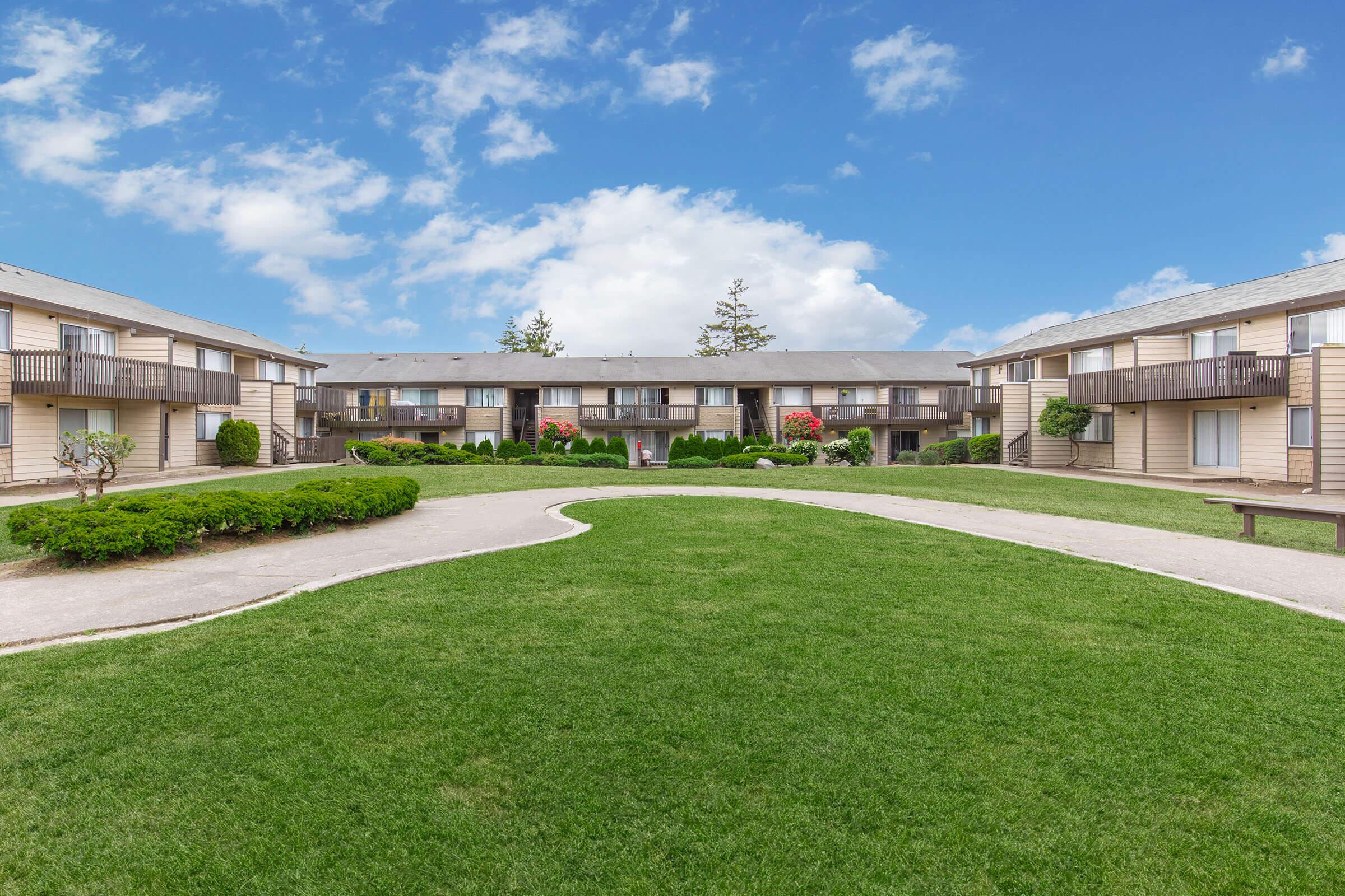 A peaceful courtyard scene featuring two-story apartment buildings surrounding a well-maintained grassy area. Lush green bushes and flowering plants add color to the landscape, under a clear blue sky with fluffy white clouds. A pathway winds through the grassy space, inviting residents to enjoy the outdoors.